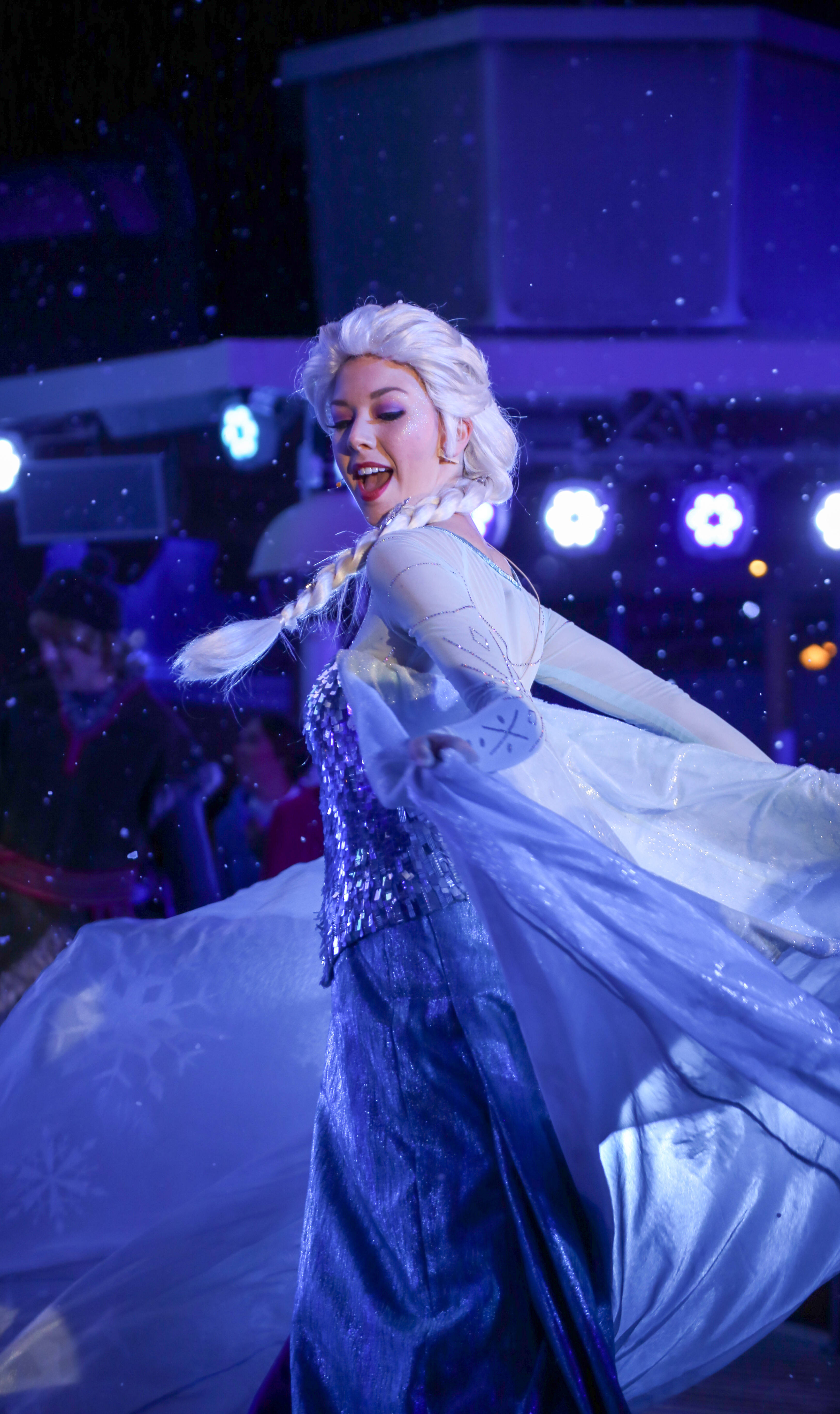 迪士尼 桌布 壁紙 — 一位扮演《冰雪奇緣》艾莎的角色扮演者在紫色舞台燈光和飄雪效果下，身穿閃亮藍色長裙旋轉。