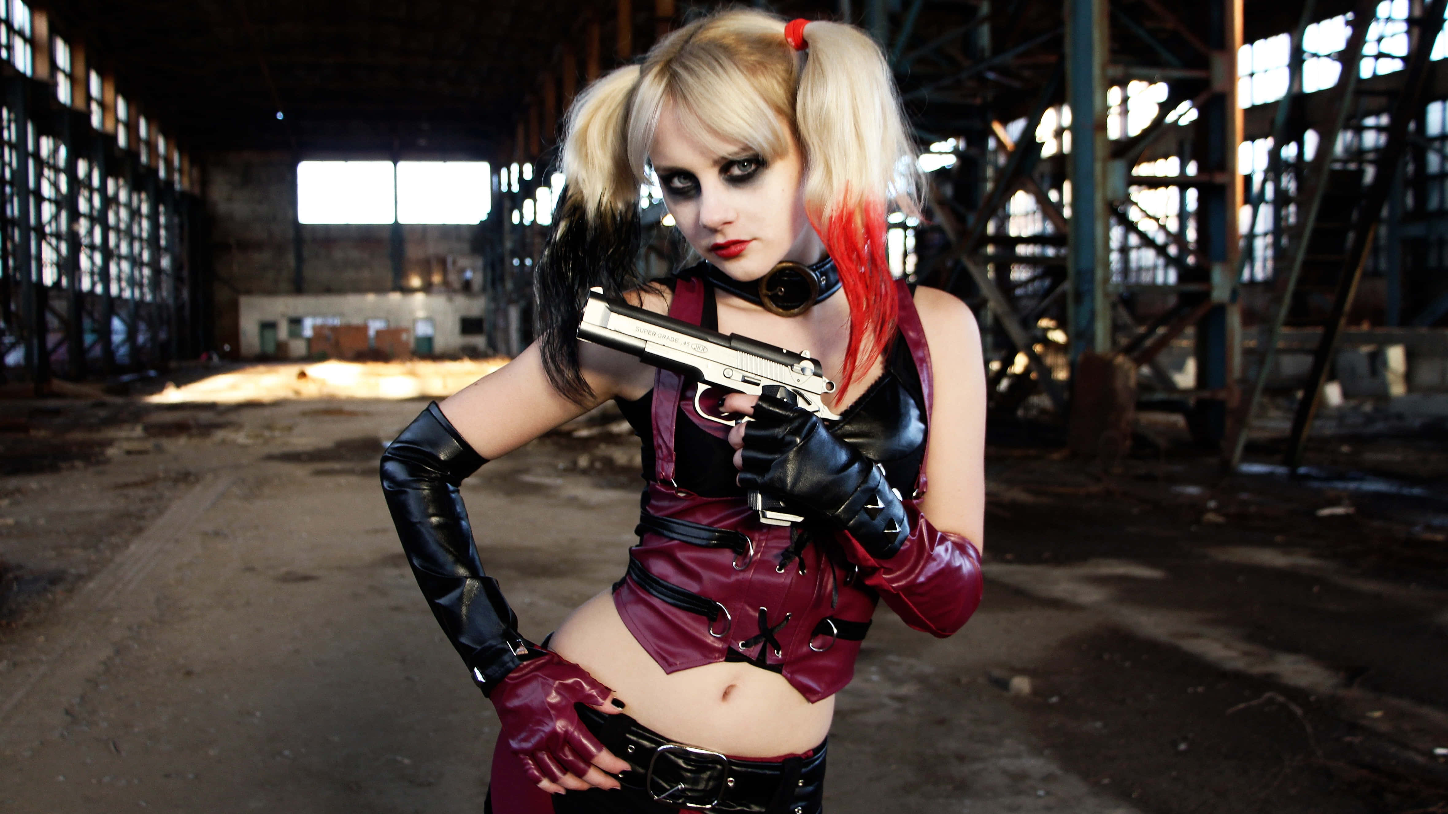 Harley Quinn Cosplay 桌布 壁紙 — 一位扮演哈莉·奎茵的角色扮演者在破舊的工業倉庫中自信地持槍擺姿勢。