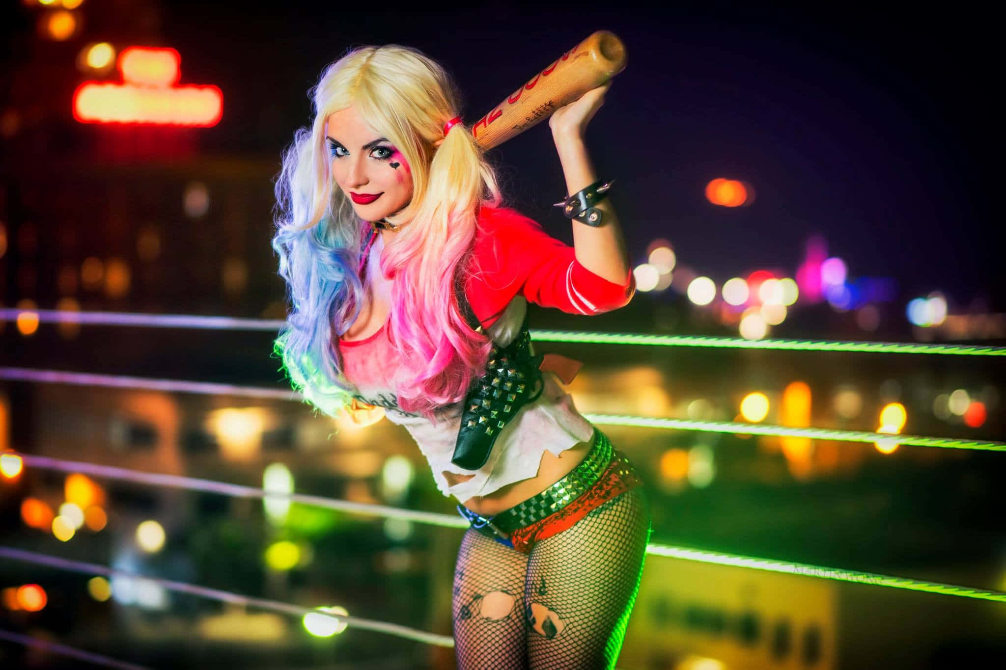 Harley Quinn Cosplay 桌布 壁紙 — 一位扮演哈莉·奎茵的角色扮演者自信地拿著棒球棍，站在摔角擂台上，背景是夜晚充滿活力的霓虹燈城市景觀。