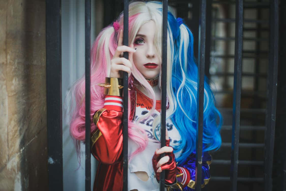 Harley Quinn Cosplay 桌布 壁紙 — 一位扮演哈莉·奎茵的角色扮演者透過黑色欄杆窺視，擁有粉紅色和藍色的頭髮以及紅唇。