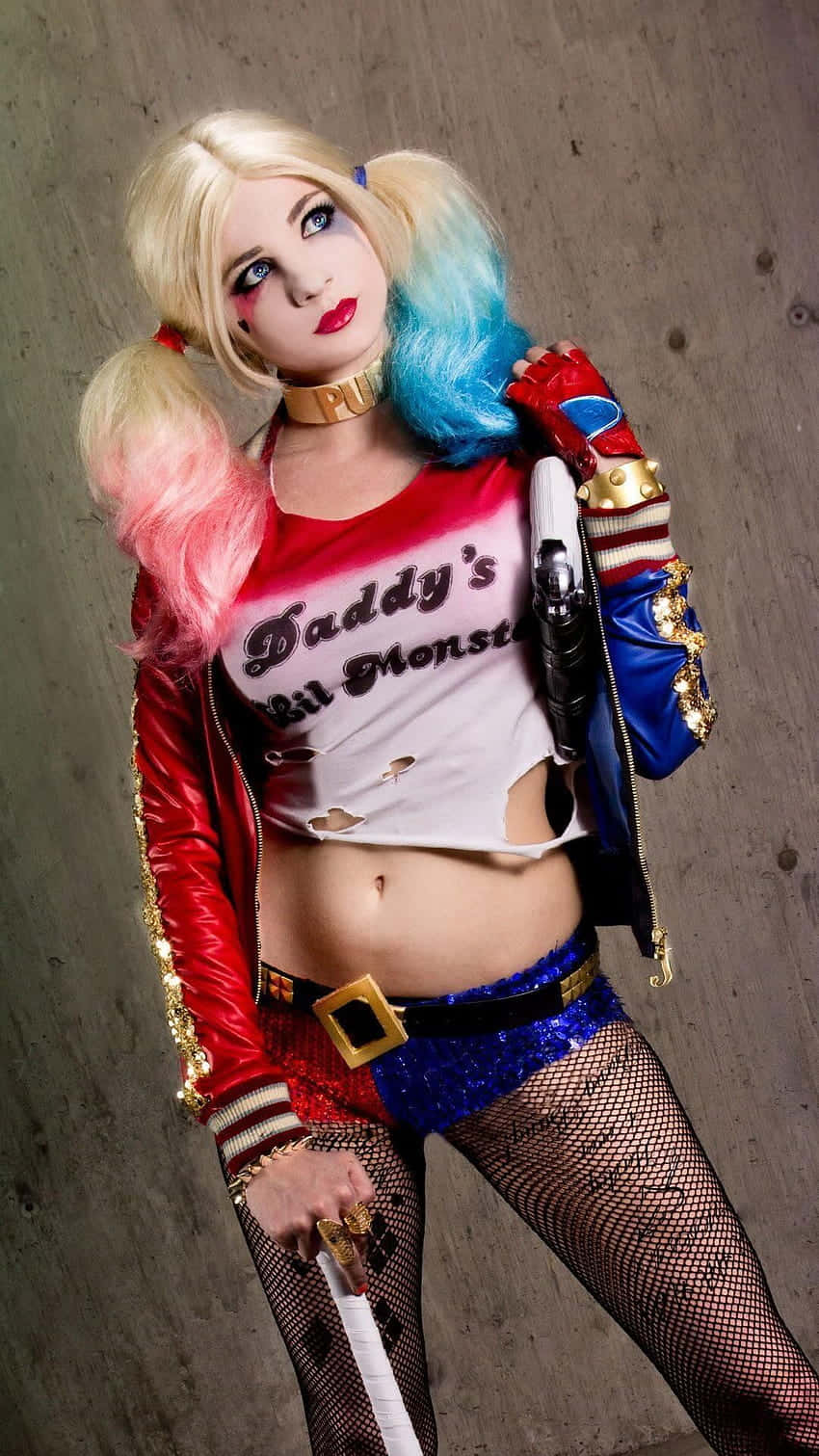 Harley Quinn Cosplay 桌布 壁紙 — 一位扮演小丑女哈莉·奎茵的cosplayer，身穿紅色外套、藍色短褲和手持槍枝，在混凝土牆前自信地擺姿勢。