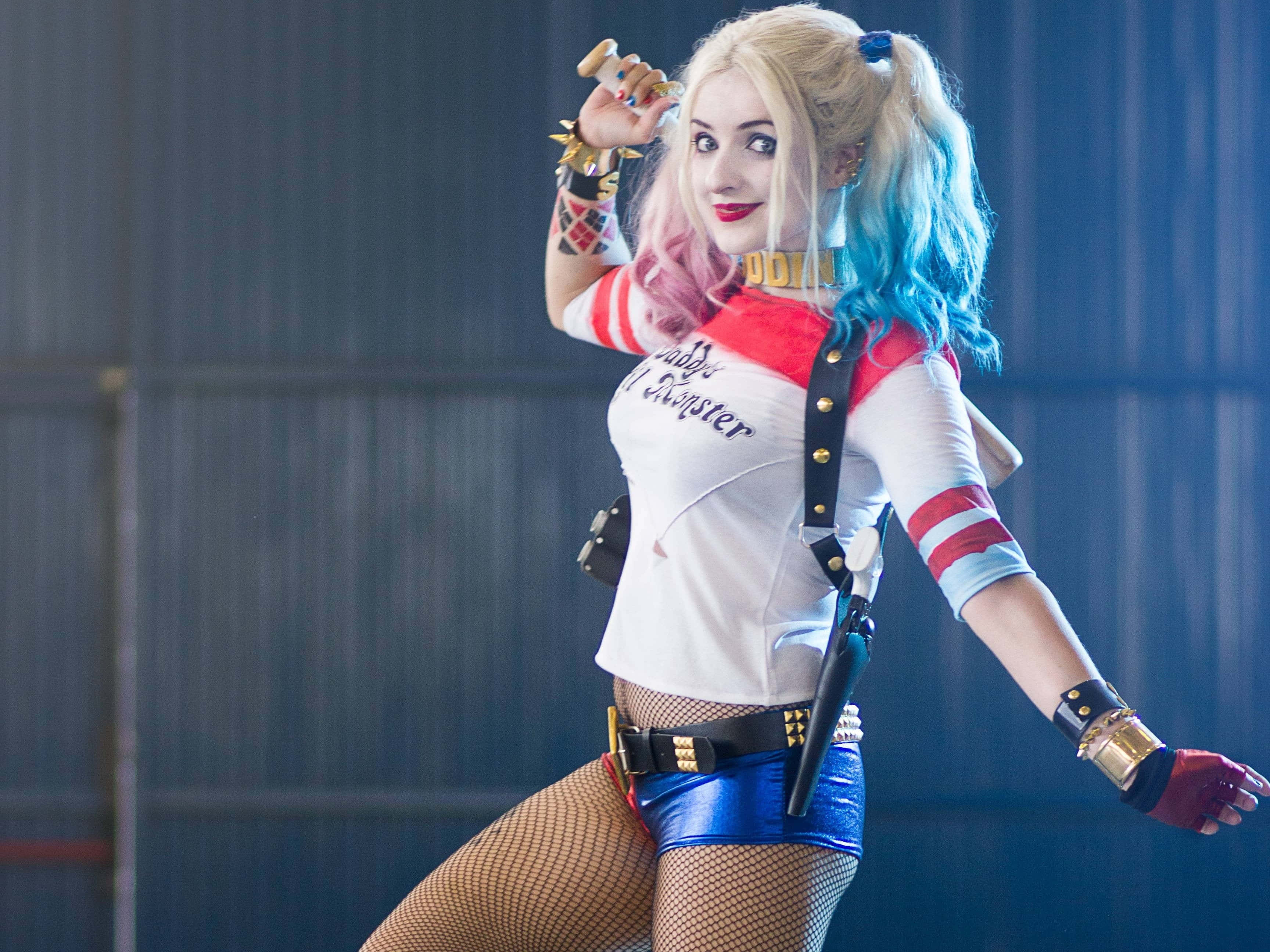 Harley Quinn Cosplay 桌布 壁紙 — 一位扮演小丑女的角色扮演者在倉庫中擺出俏皮的姿勢，擁有色彩繽紛的頭髮和標誌性服裝。