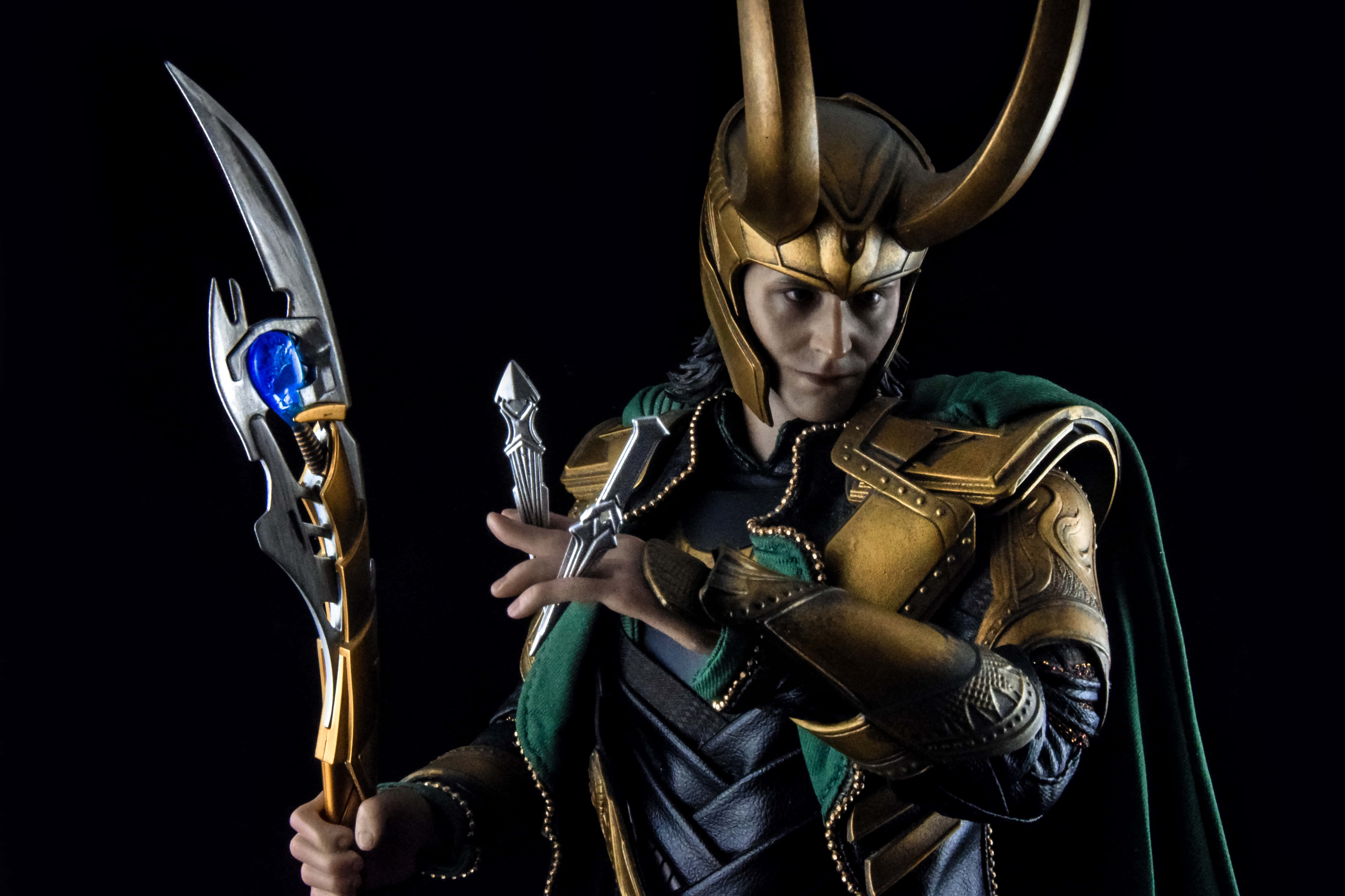 Marvel Loki 桌布 壁紙 — 一位扮演洛基的角色扮演者，在黑色背景前手持發光權杖和匕首，身穿金色盔甲和綠色斗篷。