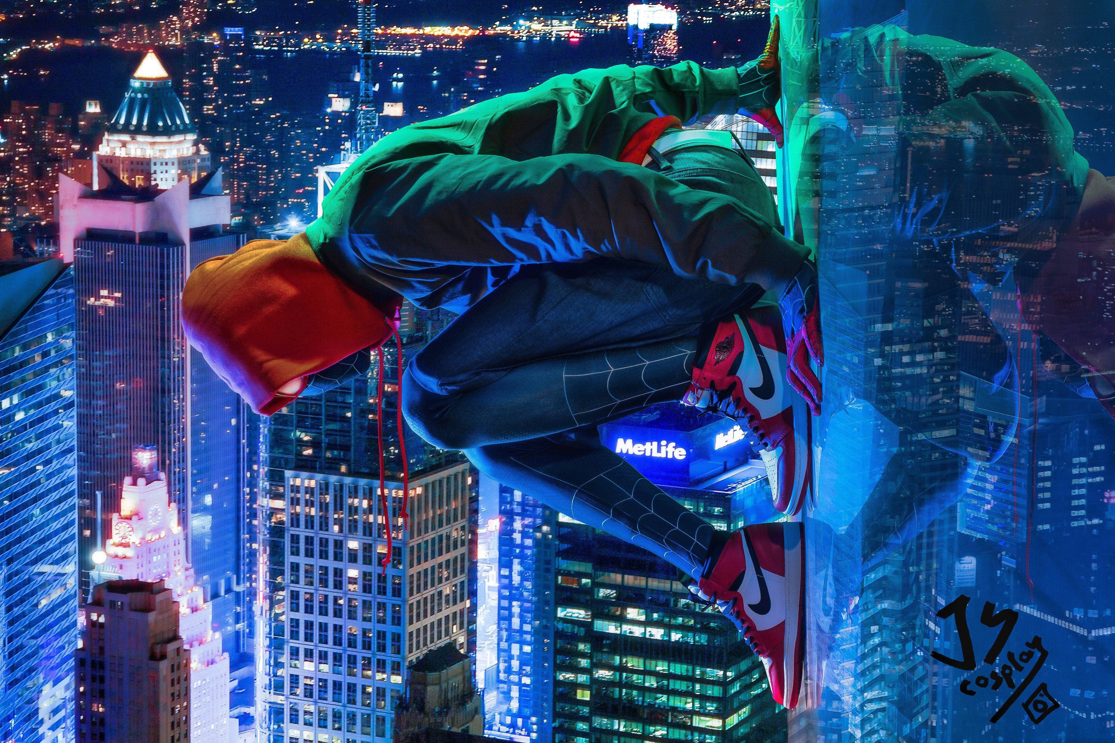 Into The Spider Verse 桌布 壁紙 — 一位扮演蜘蛛人的角色扮演者在夜間倒掛在發光的摩天大樓上，在充滿活力的城市景觀上方擺盪。