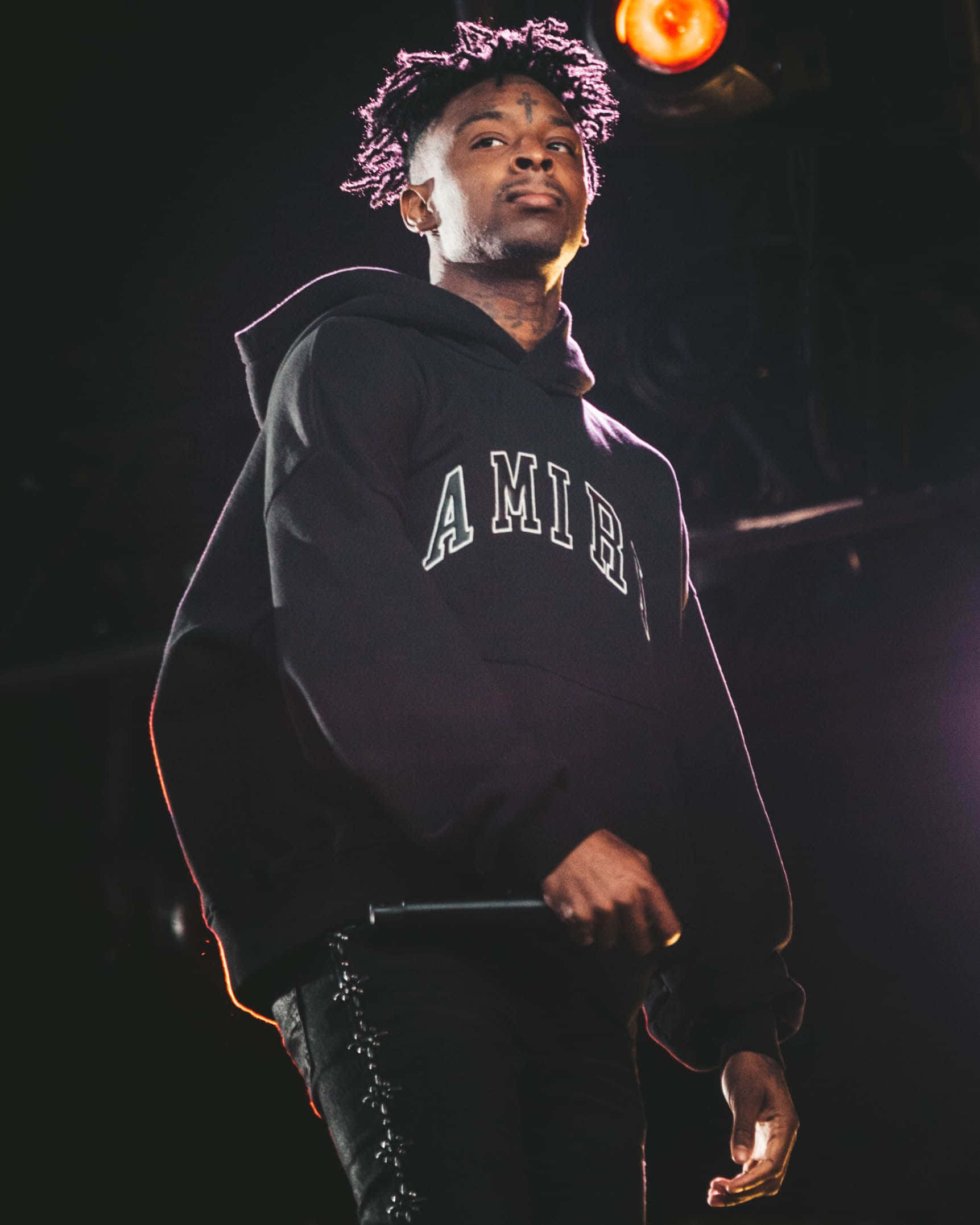 21 Savage 桌布 壁紙 — 一位捲髮、身穿連帽衫的男性表演者站在燈光昏暗的舞台上，在溫暖的聚光燈下握著麥克風。