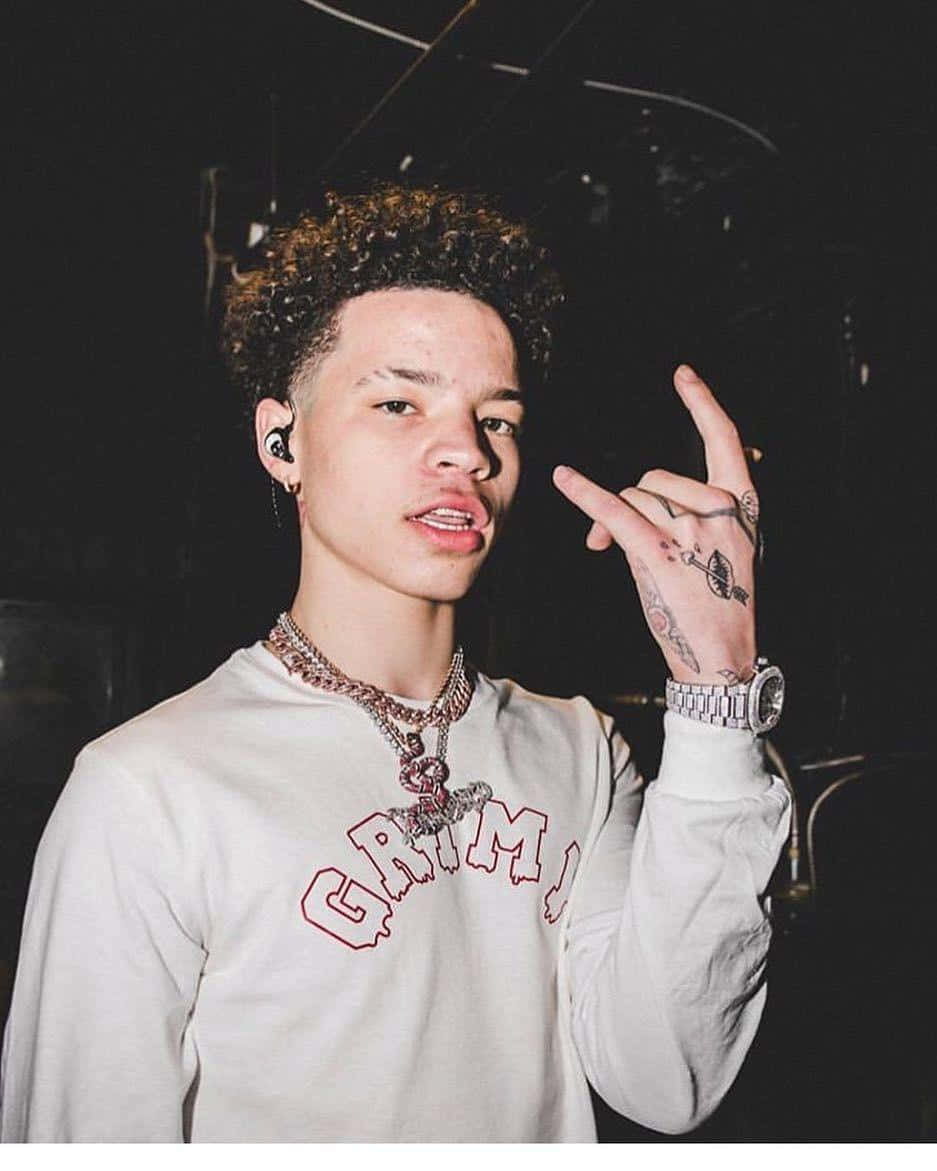 Lil Mosey 桌布 壁紙 — 一位捲髮且有刺青的年輕男子在舞台燈光下穿著白色「GRIM」連帽衫和疊戴鏈條，做出手勢。
