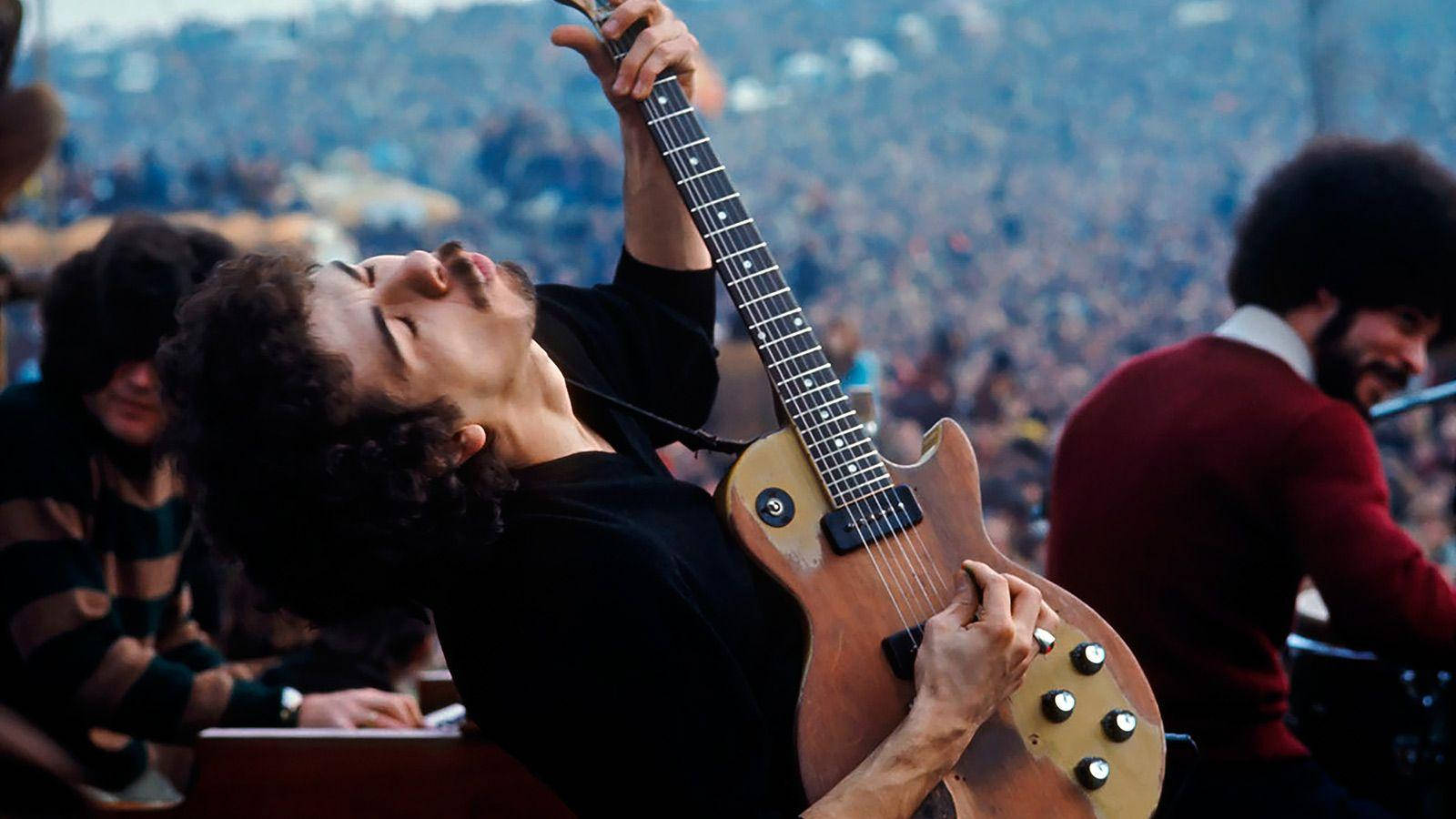 guitar player 桌布 壁紙 — 一位捲髮吉他手在舞台上對著大量觀眾熱情地彈奏一把旭日色電吉他。