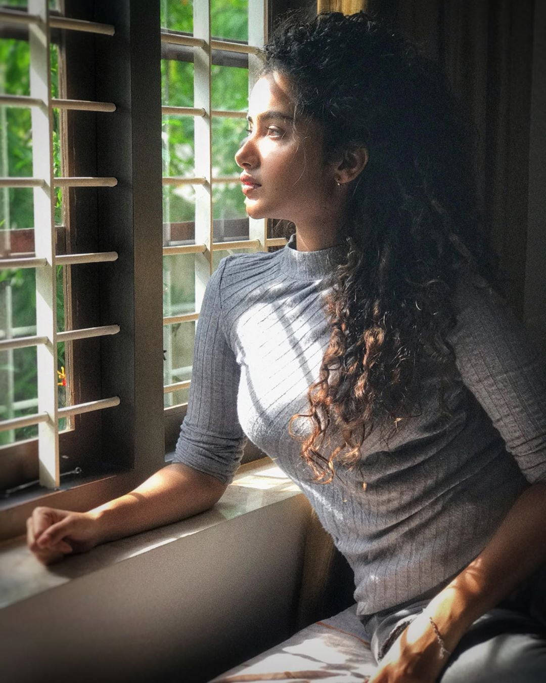 anupama parameswaran 桌布 壁紙 — 一位捲髮女士坐在陽光灑落的窗邊，靜靜地凝視窗外。