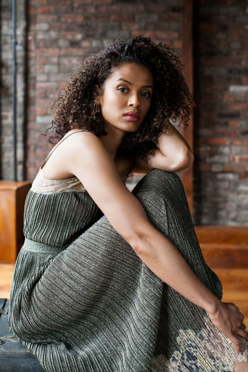 Gugu Mbatha-raw 桌布 壁紙 — 一位捲髮女士穿著質感綠色連衣裙，若有所思地坐在磚牆旁。