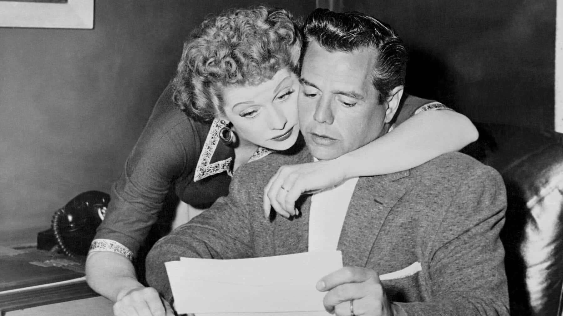 i love lucy 桌布 壁紙 — 一位捲髮女士親密地俯身靠近一位穿西裝的男士，兩人在復古室內環境中一起查看文件。