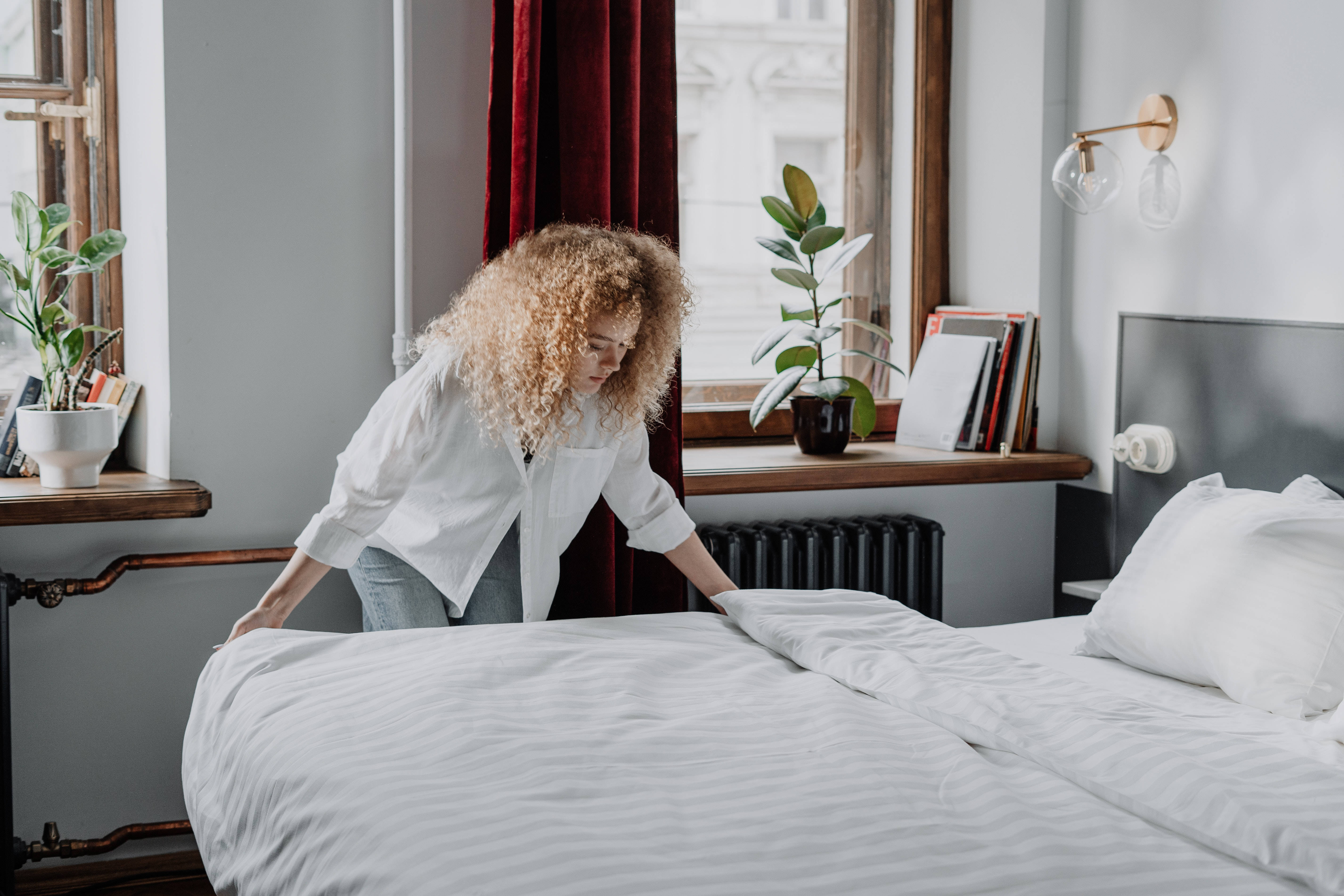 bed 桌布 壁紙 — 一位捲髮女性在一個時尚的房間裡整理床鋪，房間有紅色窗簾和盆栽植物。