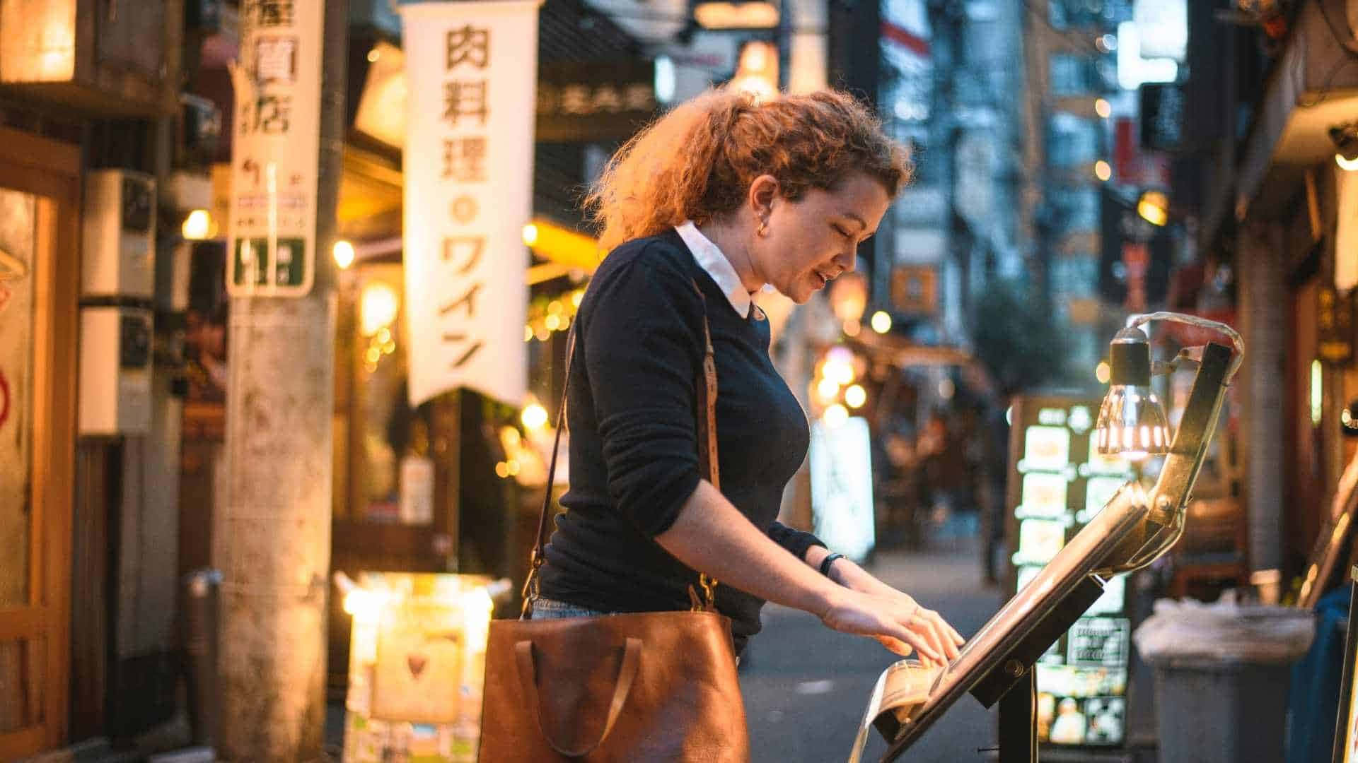 Tokyo Japan 桌布 壁紙 — 一位捲髮女性在夜晚繁忙的東京小巷中，於溫暖的霓虹燈下微笑著彈奏街頭鋼琴。