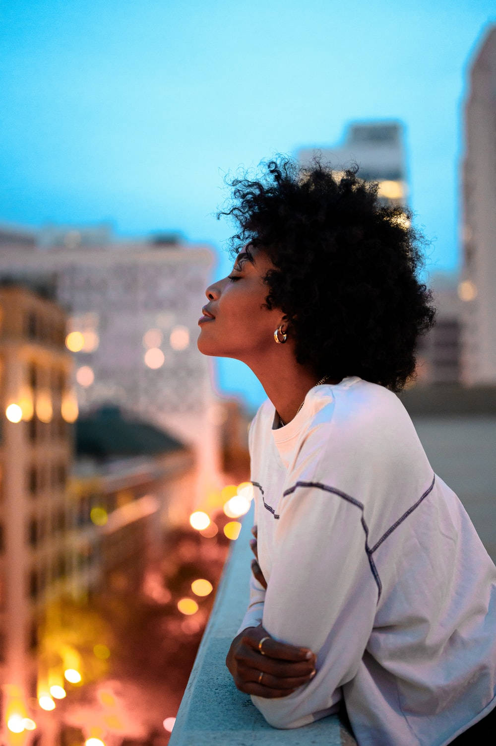 Black Girl 桌布 壁紙 — 一位捲髮女性在屋頂上仰望，城市燈光在暮色天空中模糊成溫暖的散景。