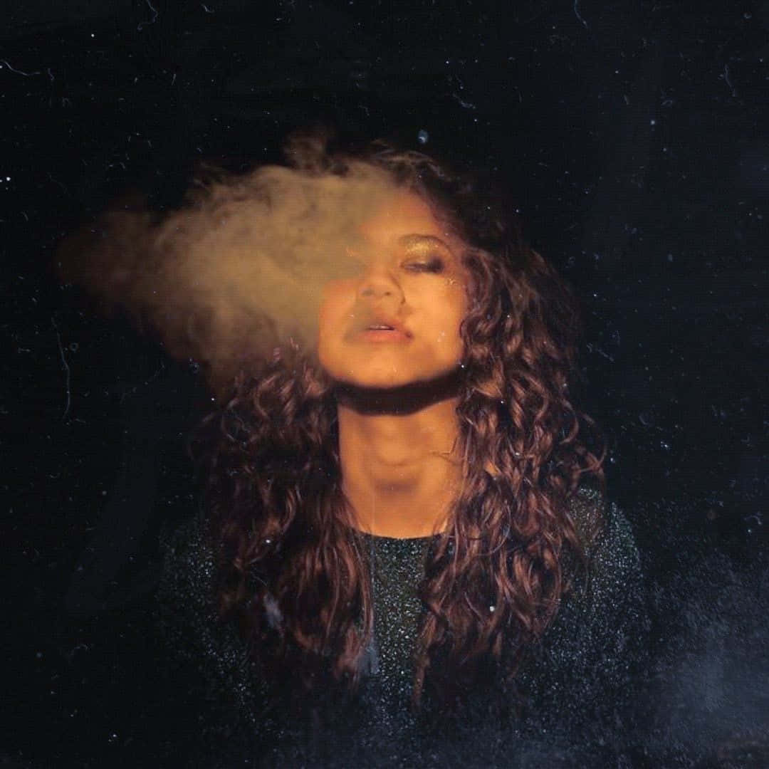 愉悅 桌布 壁紙 — 一位捲髮女性在昏暗星空背景下，柔和燈光中呼出一縷煙霧。
