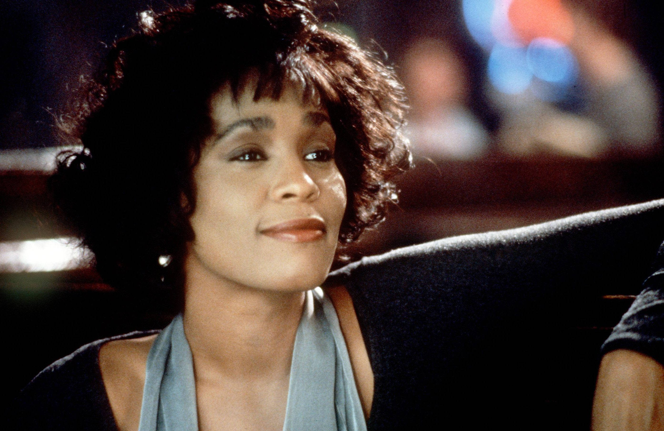 Whitney 桌布 壁紙 — 一位捲髮女性在昏暗的房間裡靠著深色沙發，身後模糊的彩色燈光中溫柔微笑。