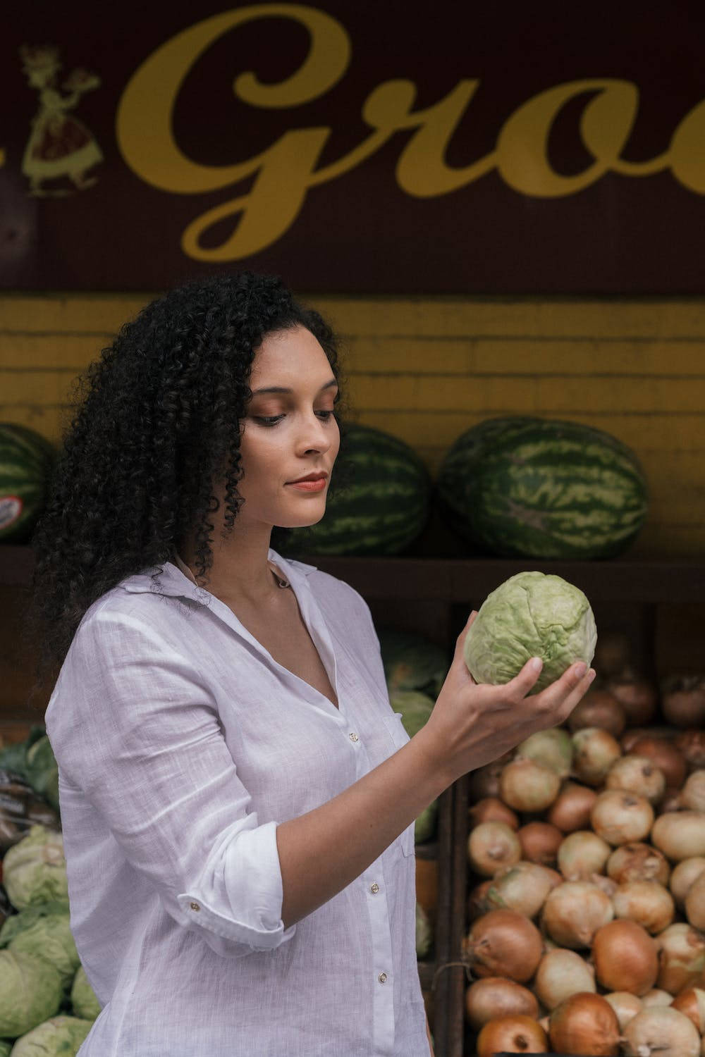 Vegetables 桌布 壁紙 — 一位捲髮女性在溫暖燈光下，於一個擺滿西瓜和洋蔥的市場攤位前拿著一顆高麗菜。