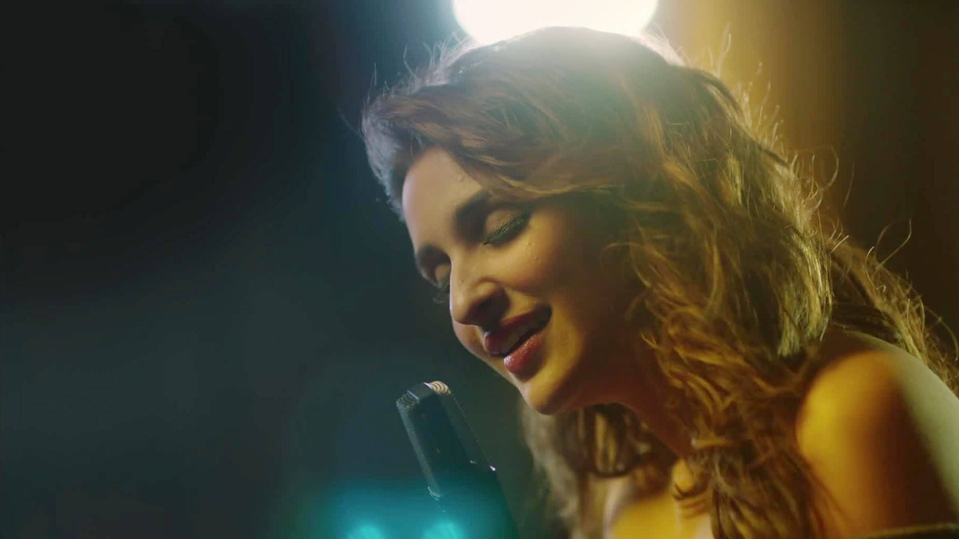 Parineeti 桌布 壁紙 — 一位捲髮女性在溫暖的舞台燈光下對著麥克風唱歌，溫柔地微笑。