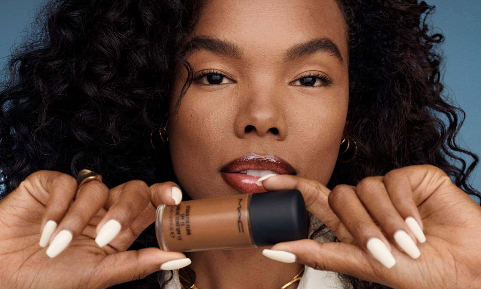 Mac Cosmetics 桌布 壁紙 — 一位捲髮女性在藍色背景前將MAC粉底液瓶靠近嘴唇。