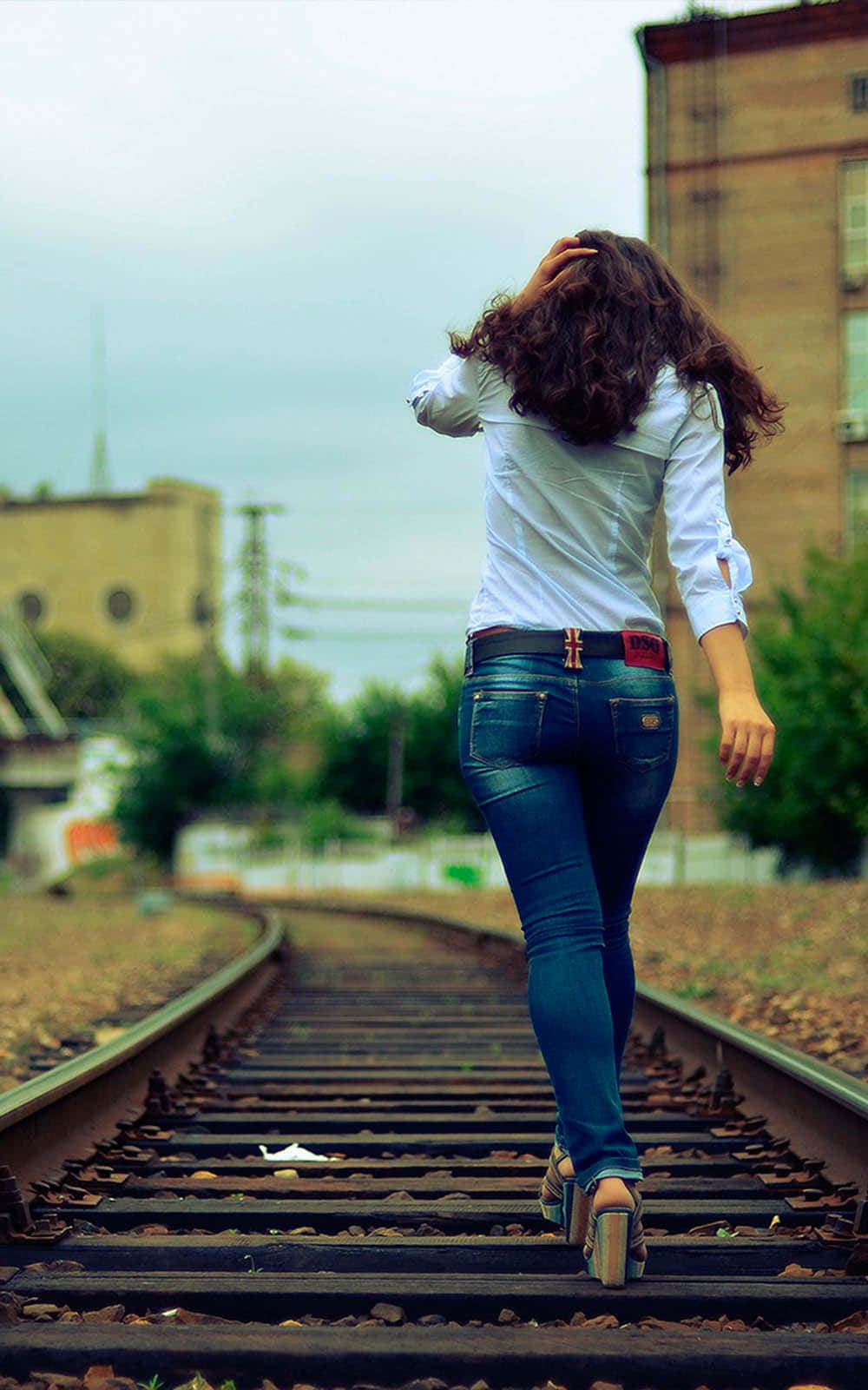 Woman 桌布 壁紙 — 一位捲髮女性在陰天下的城市景觀中，沿著風化的火車軌道走遠。
