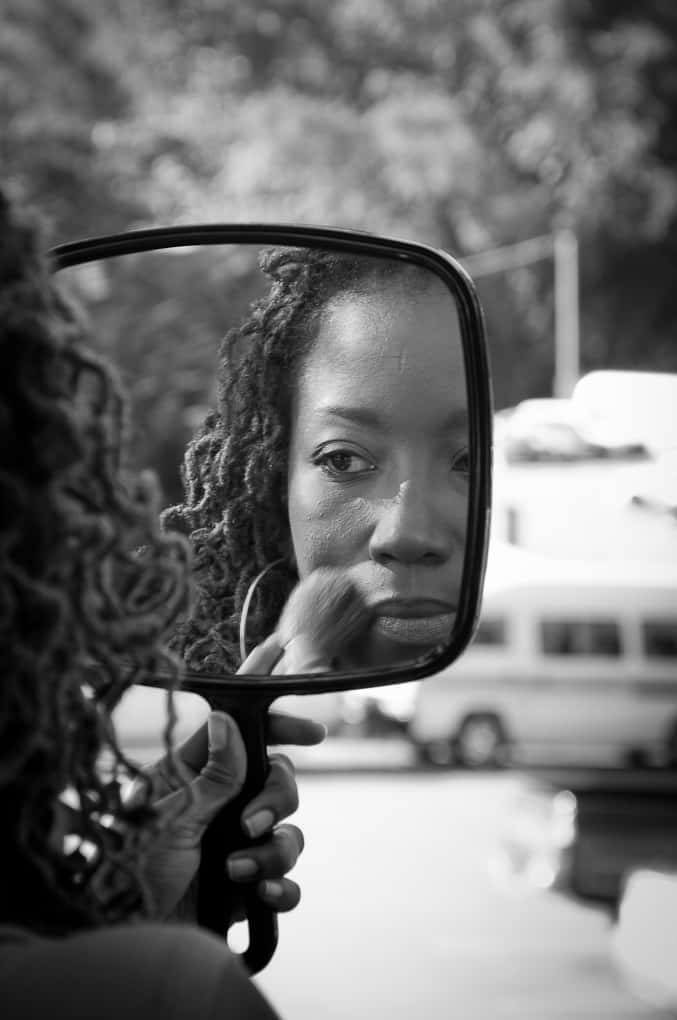 reflection 桌布 壁紙 — 一位捲髮女性坐在車內，手持鏡子化妝，黑白拍攝，背景為模糊的戶外景色。