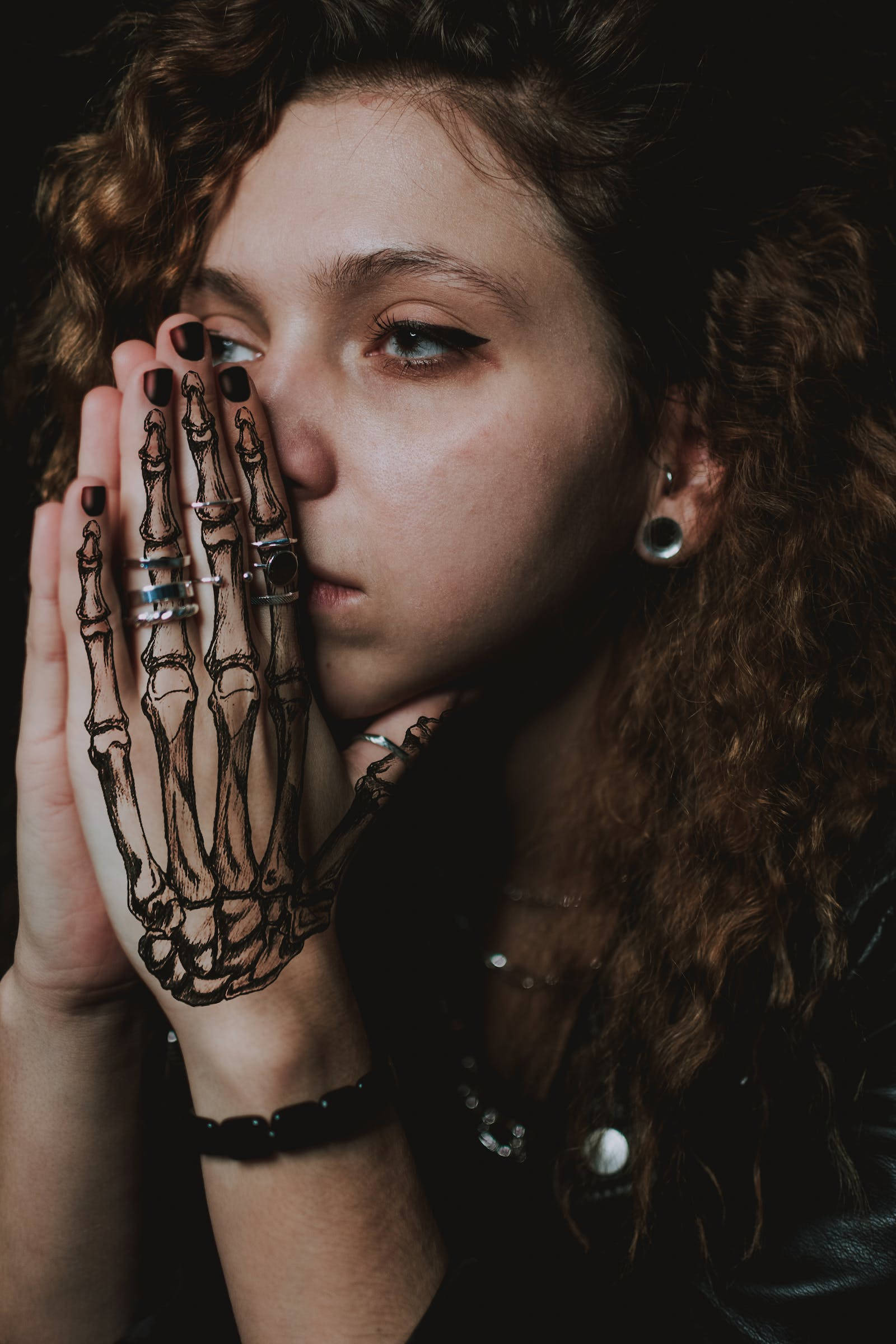 Cool Sad 桌布 壁紙 — 一位捲髮女性將手靠近臉部，在戲劇性燈光下露出詳細的骷髏手部刺青。