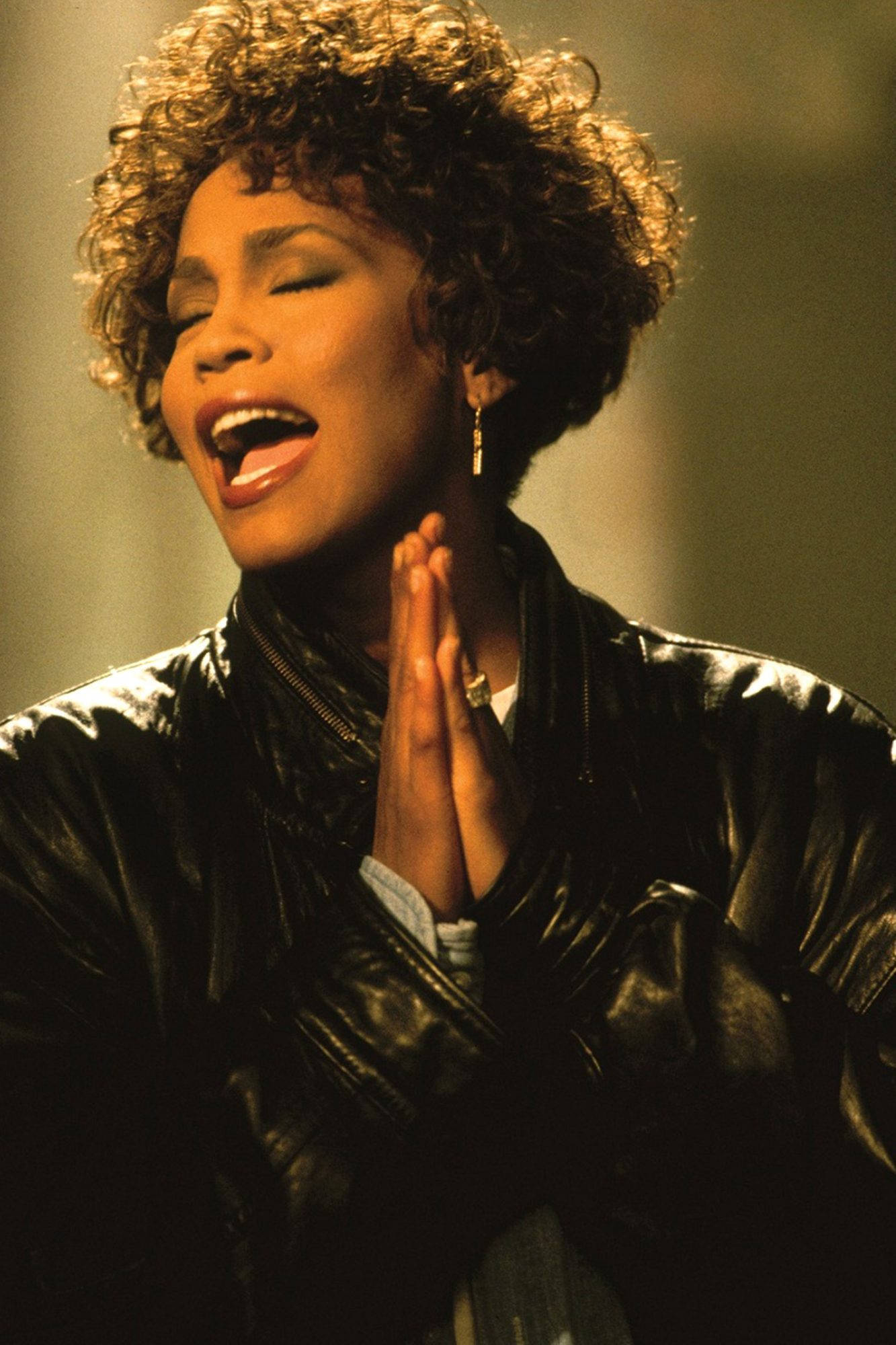 Whitney 桌布 壁紙 — 一位捲髮女性穿著黑色皮夾克，雙手交疊在胸前，熱情地唱歌。