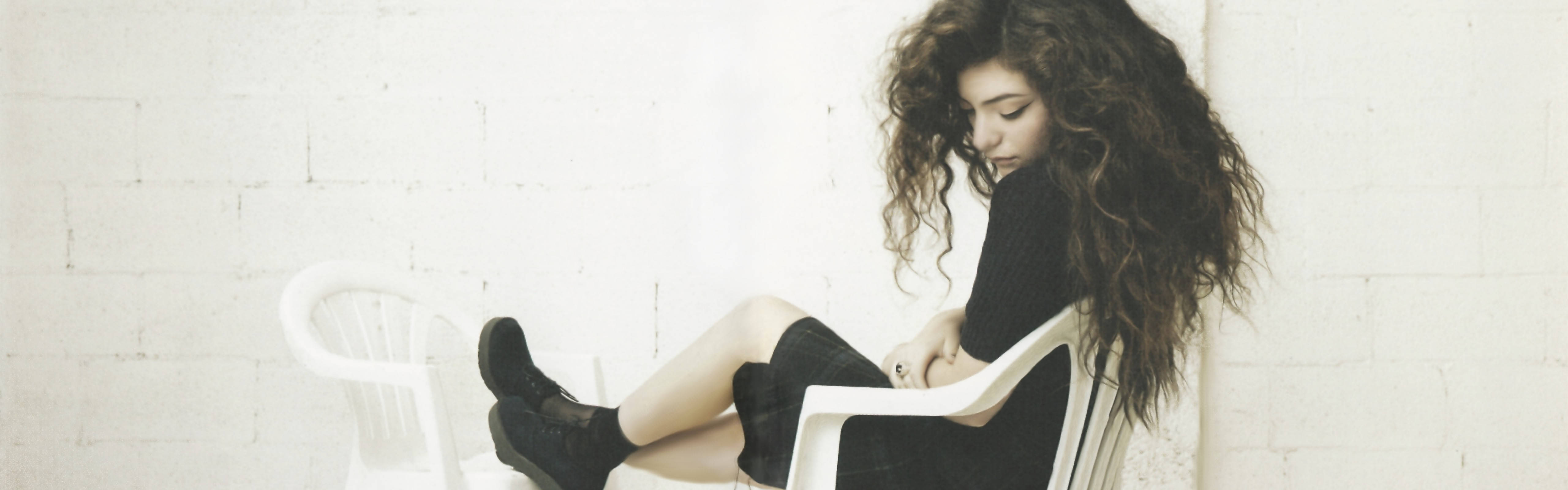 Lorde 桌布 壁紙 — 一位捲髮女性穿著黑色衣服，坐在白色塑膠椅上，靠著白色磚牆沉思。