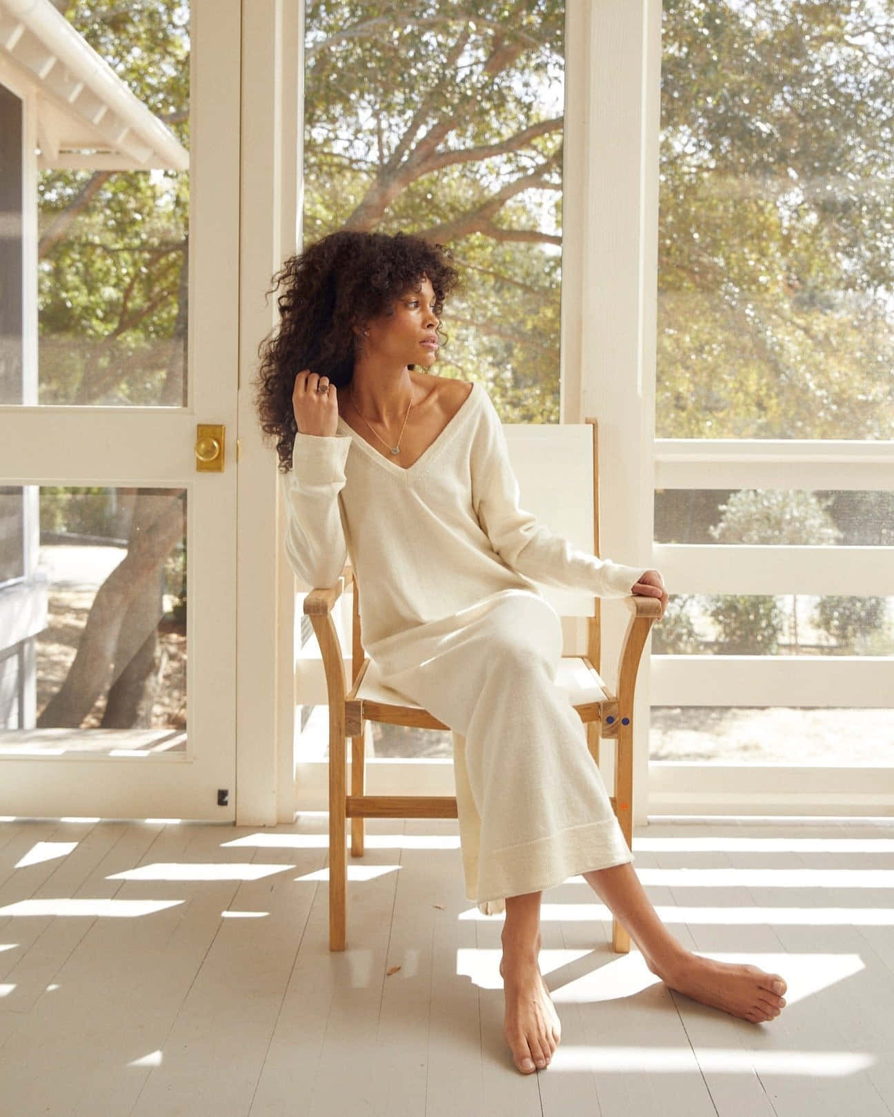 Minimalist Fashion 桌布 壁紙 — 一位捲髮女性赤腳穿著奶油色針織連衣裙坐在木椅上，溫暖的陽光穿過玻璃門灑在白色木地板上。