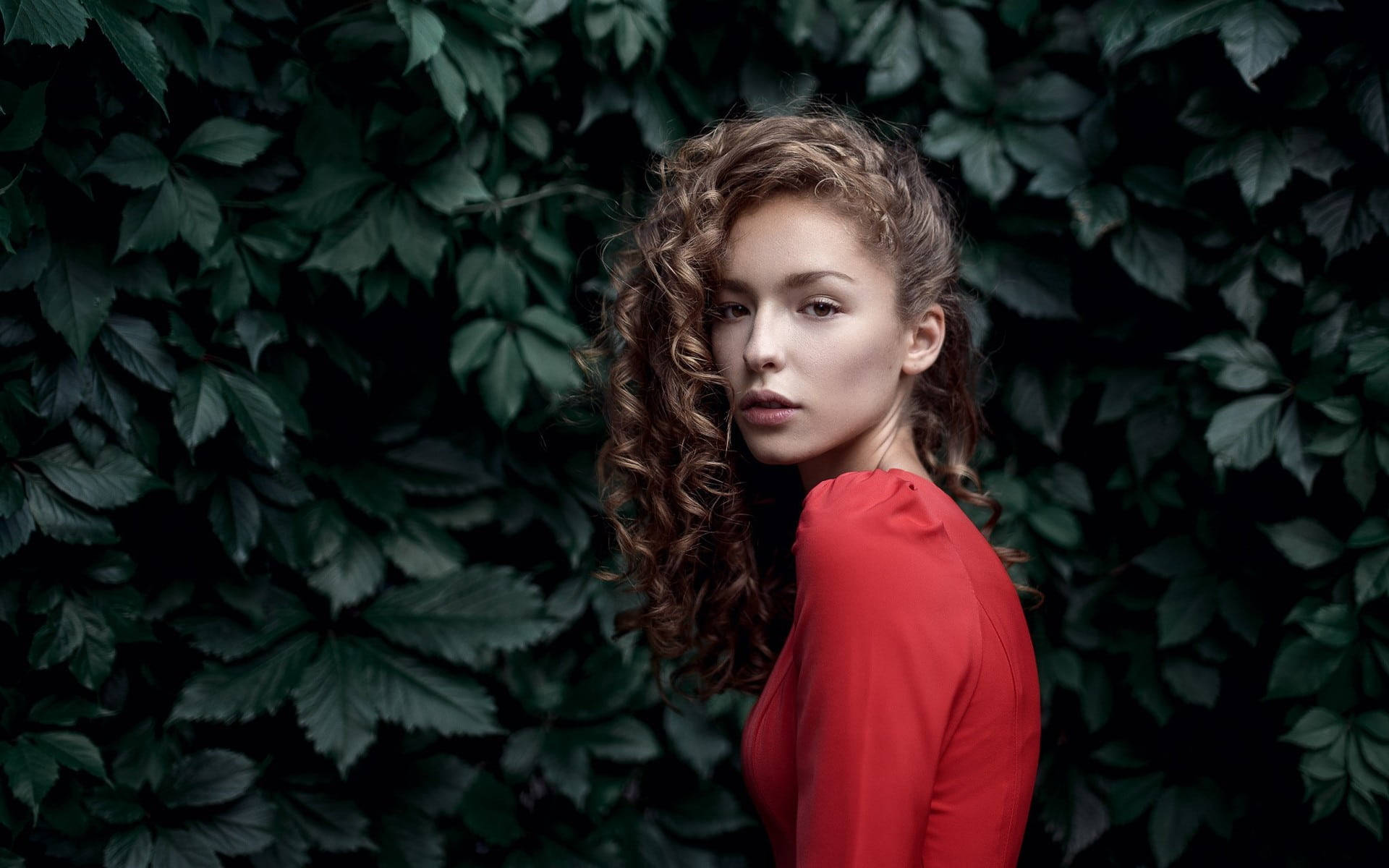 Beautiful Woman 桌布 壁紙 — 一位捲髮女性身穿紅色上衣，靠著濃密的深綠色葉牆，回頭望向肩膀後方。