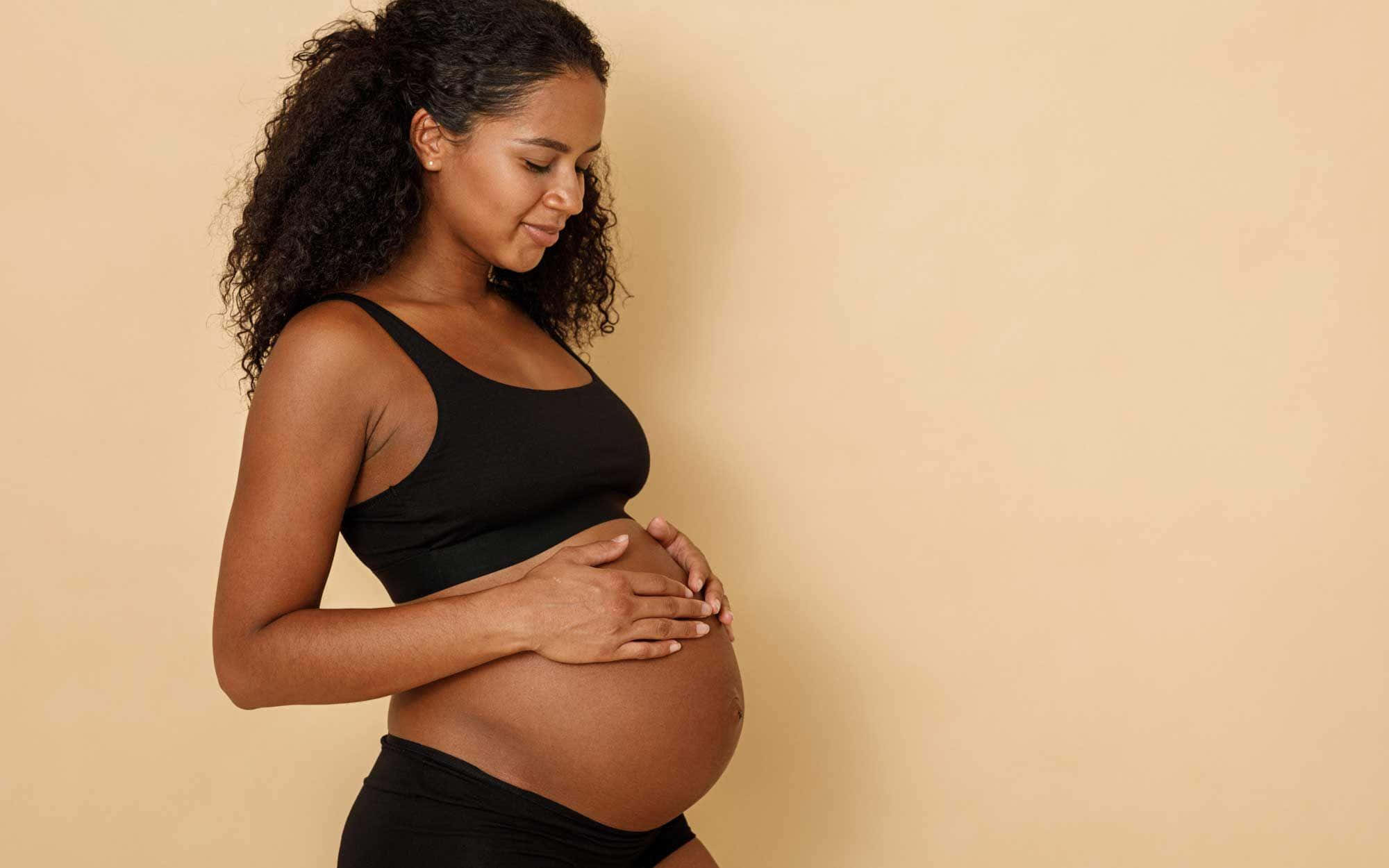 Pregnant 桌布 壁紙 — 一位捲髮孕婦輕柔地抱著肚子，靠在柔軟的米色牆上，展現溫柔一刻。