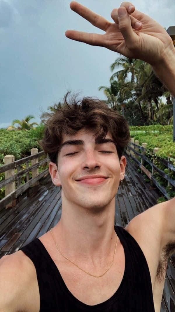 美學 桌布 壁紙 — 一位捲髮年輕男子在木橋上閉眼微笑，周圍環繞著茂密的綠植和棕櫚樹。