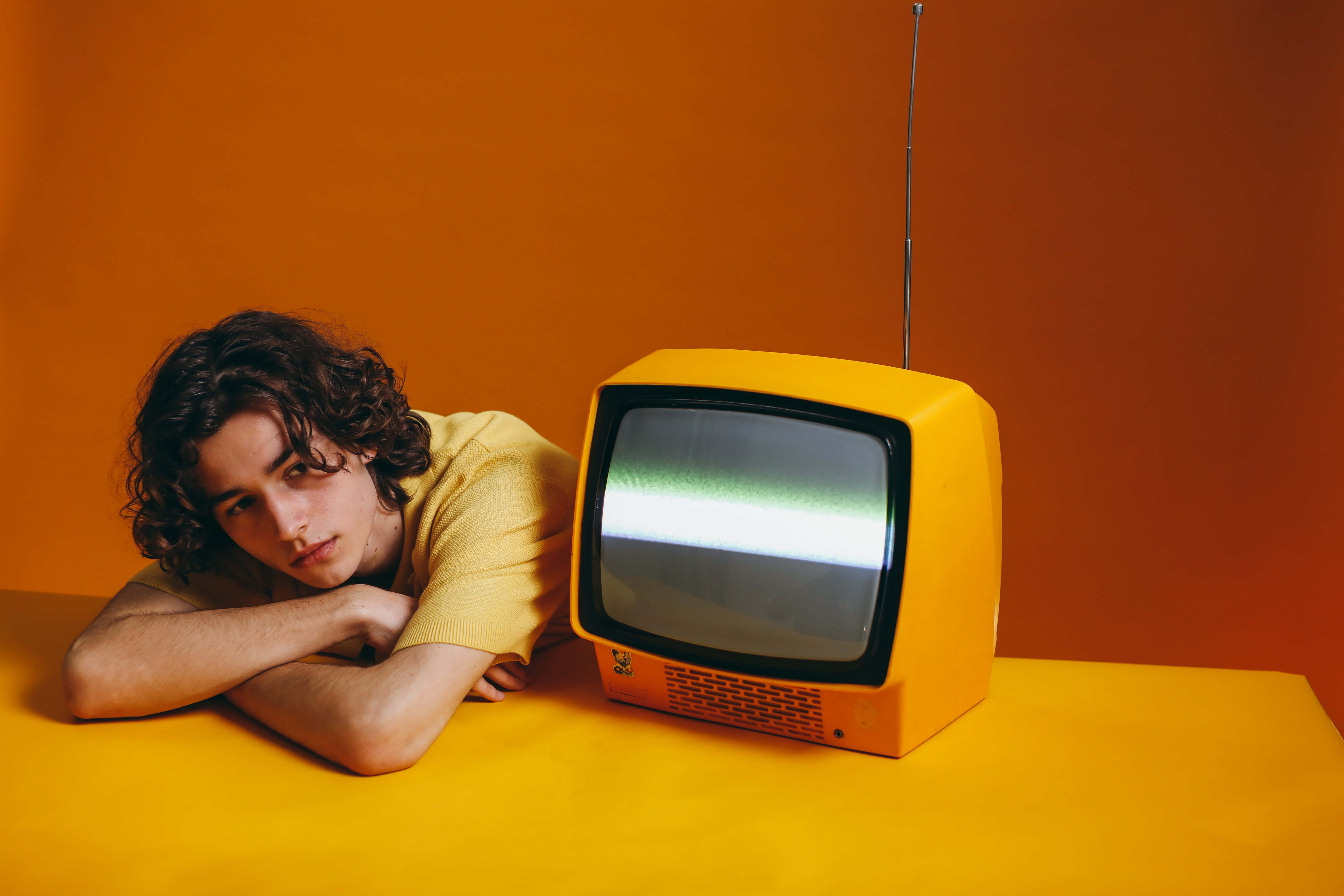 television 桌布 壁紙 — 一位捲髮年輕男子在溫暖的橙色背景下，將頭靠在手臂上，旁邊是一台發光的復古黃色電視機。