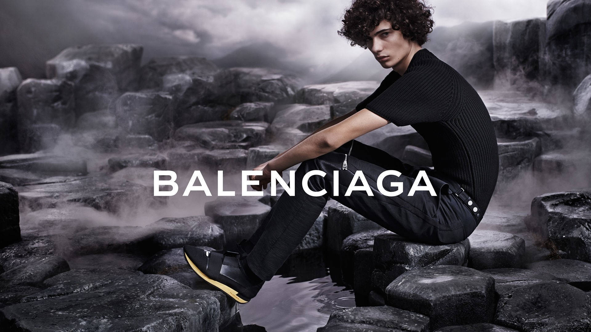 Balenciaga 桌布 壁紙 — 一位捲髮年輕男子坐在霧氣瀰漫的火山岩上，身穿黑色 Balenciaga 服裝和黃色鞋底的鞋子。