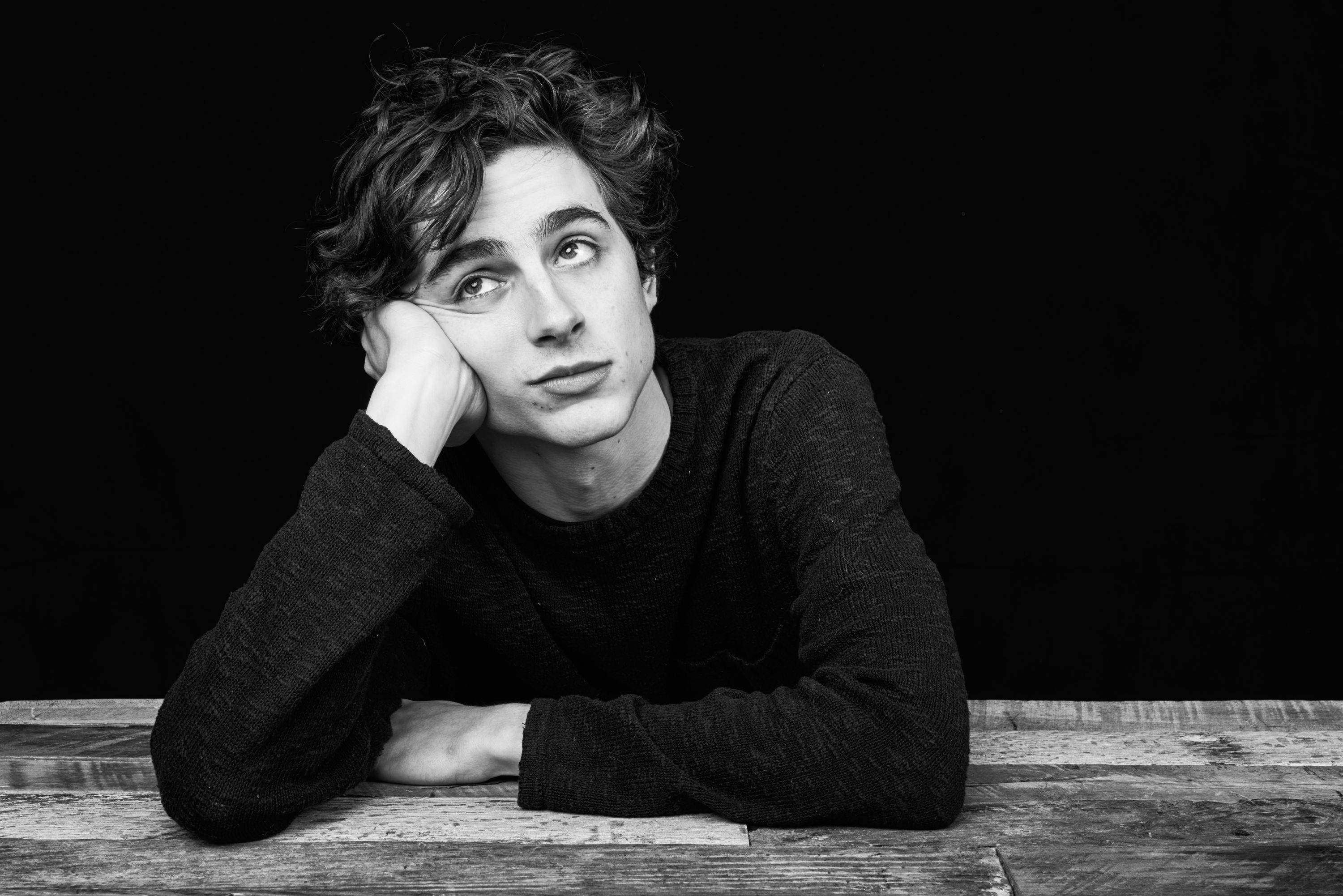 Timothée Chalamet 桌布 壁紙 — 一位捲髮年輕男子將下巴擱在手上，對著黑色背景沉思地向上凝視。