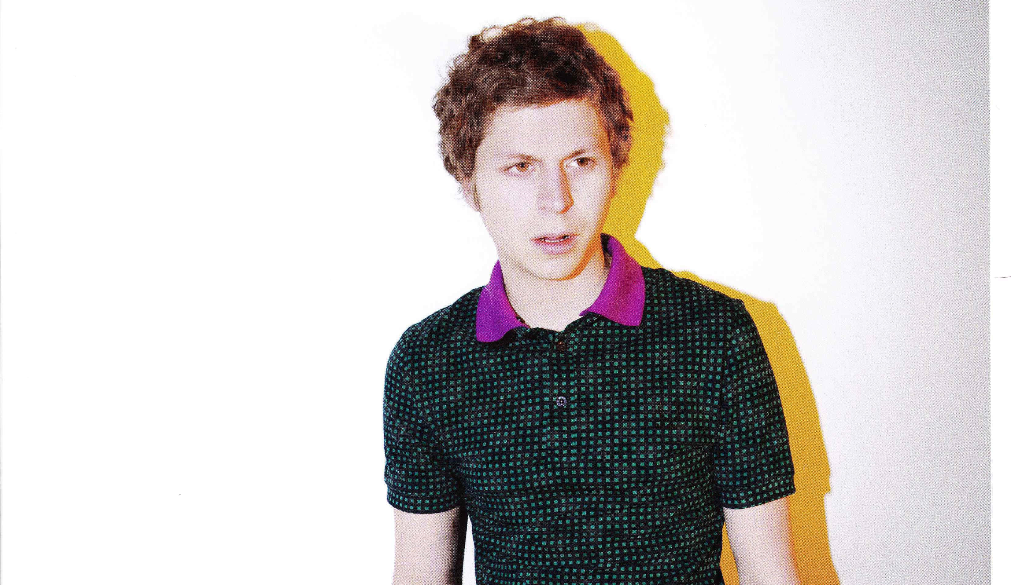 michael cera 桌布 壁紙 — 一位捲髮年輕男子穿著深綠色圓點Polo衫，配紫色衣領，站在純白牆壁前，投射出大膽的黃色陰影。