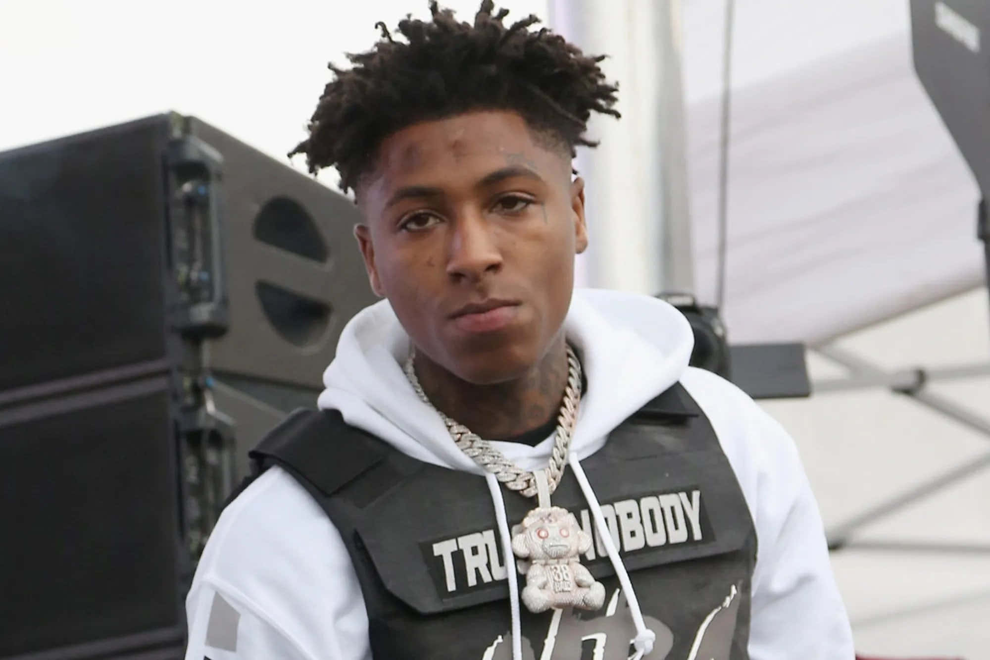 NBA YoungBoy 桌布 壁紙 — 一位捲髮年輕男子穿著白色連帽衫，外搭黑色背心，上面有'TRU NOBODY'文字和泰迪熊吊墜項鍊。