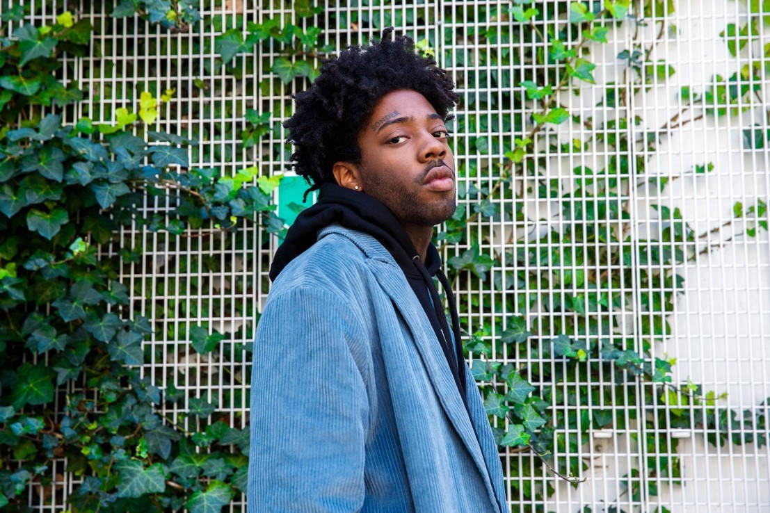 Brent Faiyaz 桌布 壁紙 — 一位捲髮年輕男子身穿藍色燈芯絨外套，自信地站在被茂密綠色常春藤覆蓋的白色網格牆前。