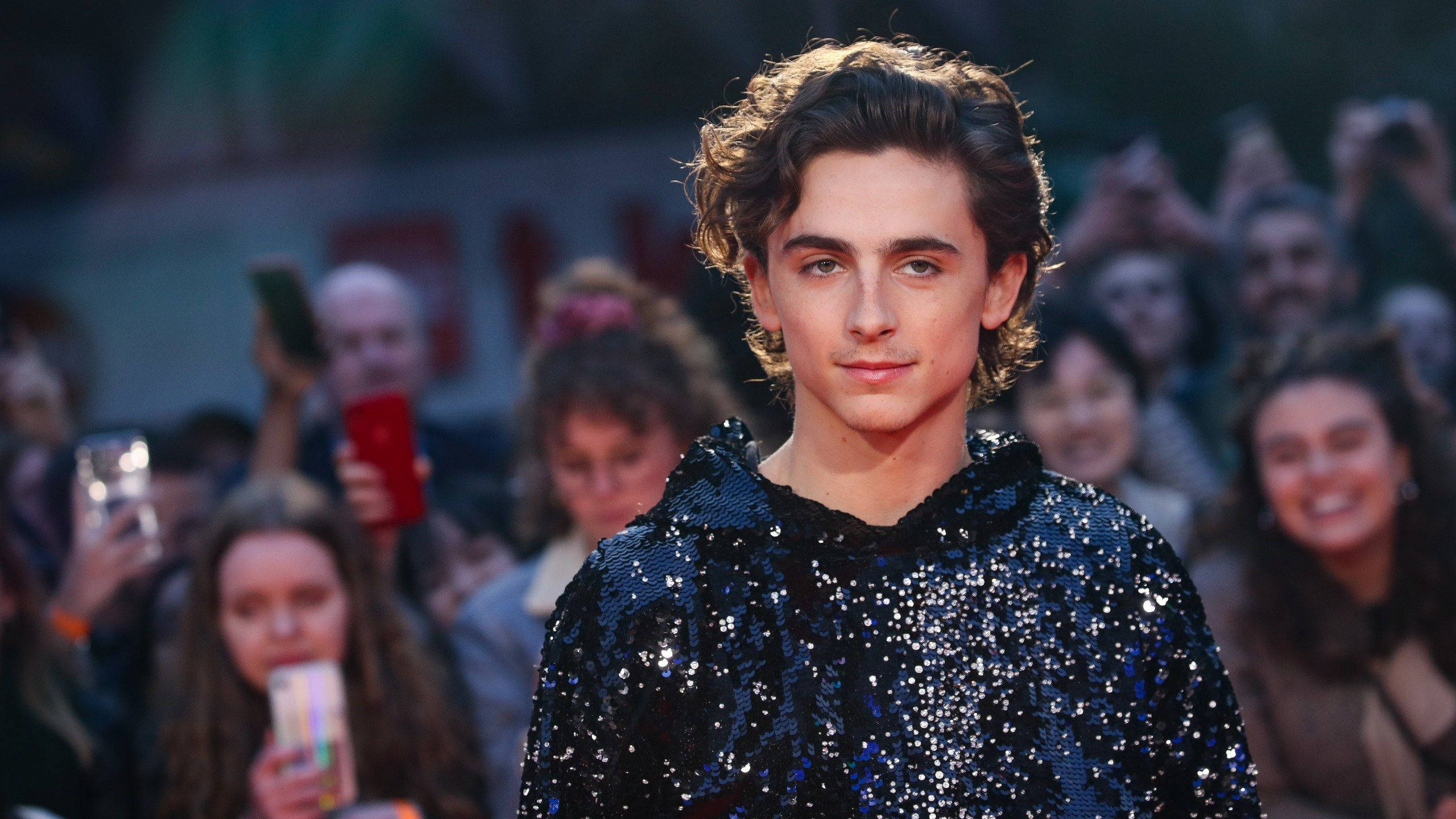 Timothée Chalamet 桌布 壁紙 — 一位捲髮年輕男子身穿閃亮藍色亮片上衣，自信地走在紅地毯上，身後粉絲舉著手機。