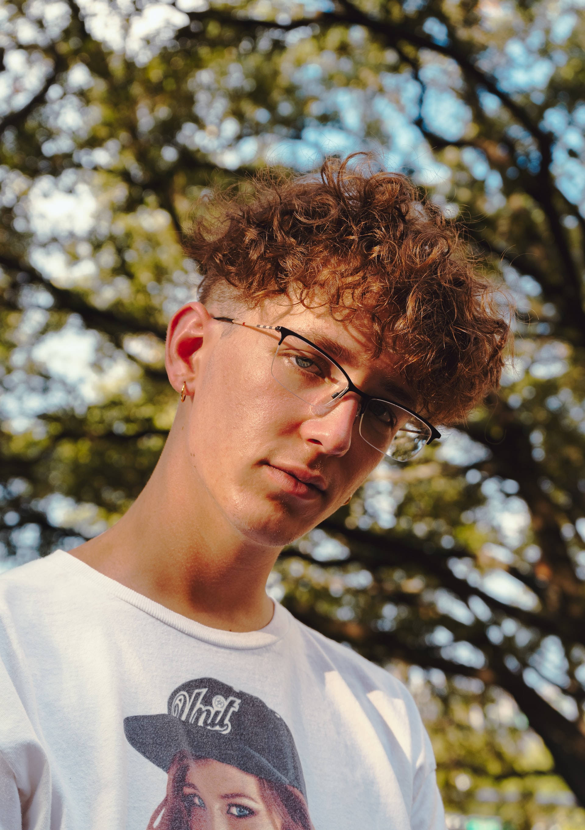 Haircut 桌布 壁紙 — 一位捲髮戴眼鏡的年輕男子在陽光照射的樹木背景下低頭注視。