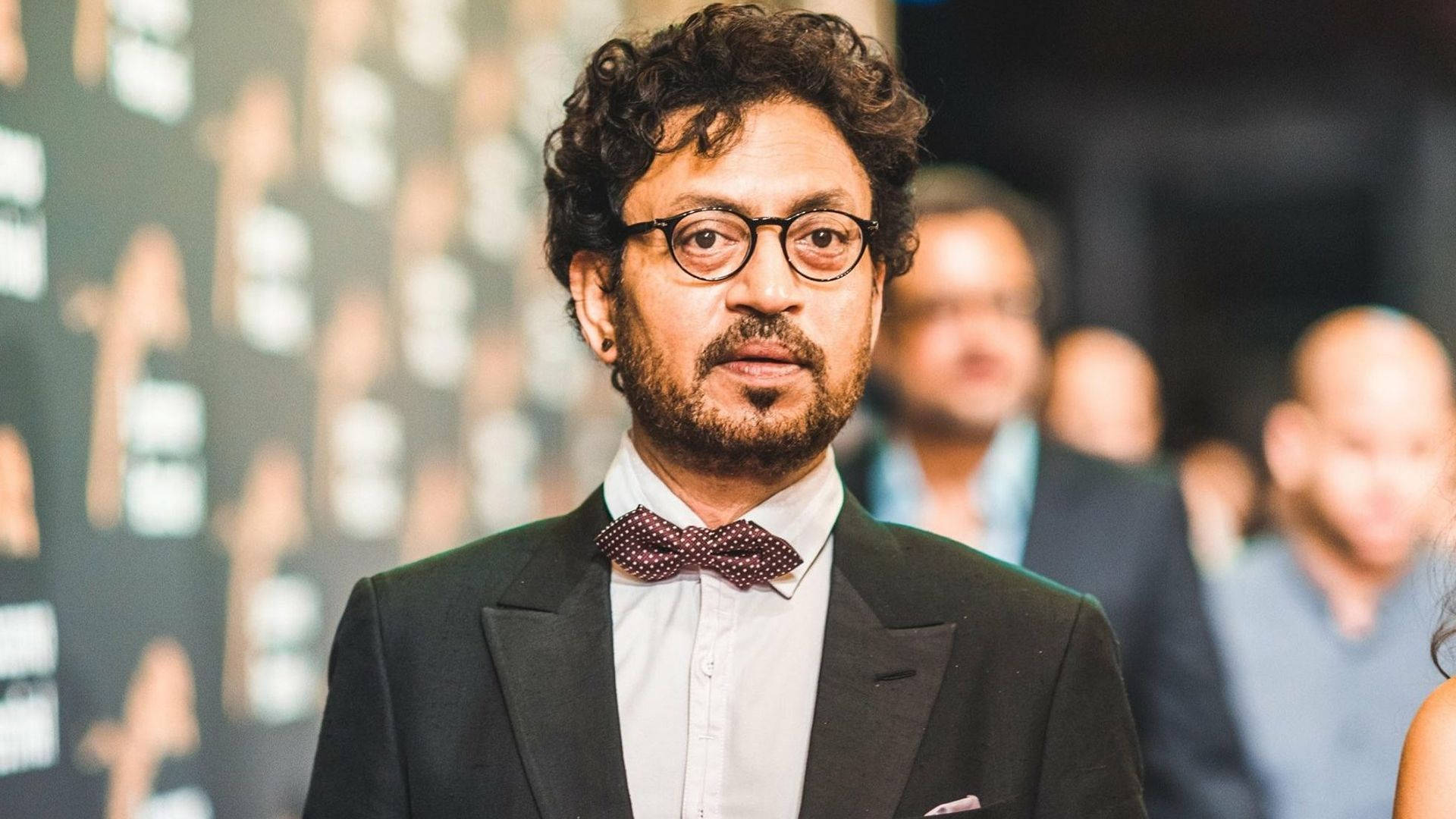 Irrfan Khan 桌布 壁紙 — 一位捲髮戴眼鏡的男子在正式場合穿著黑色西裝和波點領結。