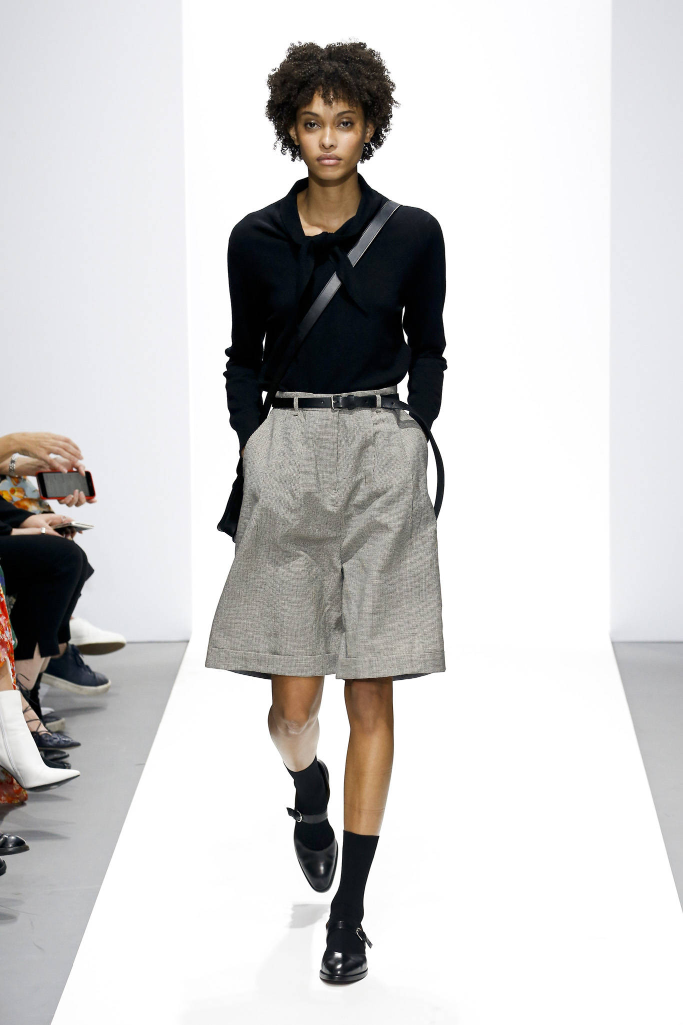 Fashion Model 桌布 壁紙 — 一位捲髮模特兒自信地走在明亮的白色天橋上，身穿黑色上衣、灰色短褲和黑色鞋子。