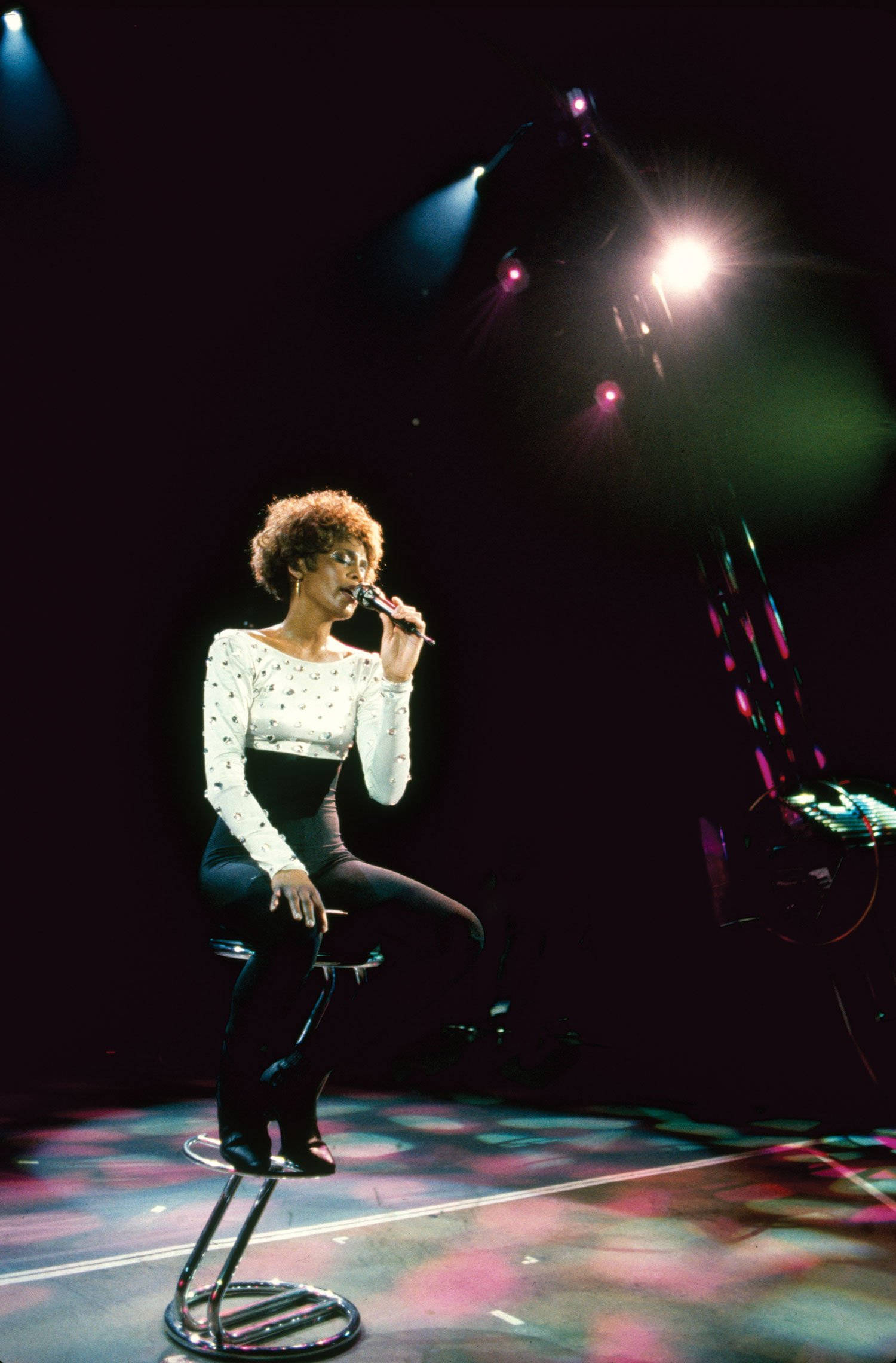 Whitney 桌布 壁紙 — 一位捲髮歌手坐在凳子上，在明亮的聚光燈下表演，舞台地板反射出繽紛色彩。