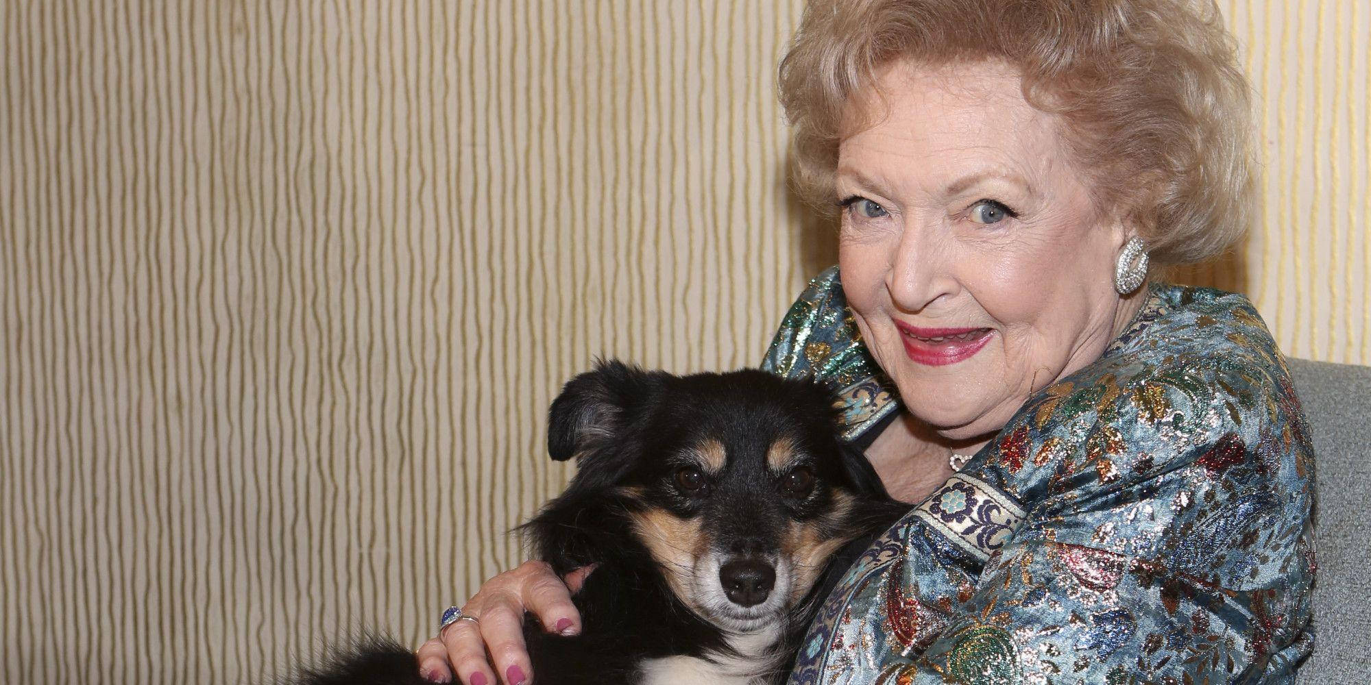 Betty White 桌布 壁紙 — 一位捲髮老婦人微笑著，在條紋壁紙背景下擁抱一隻小黑褐色小狗。