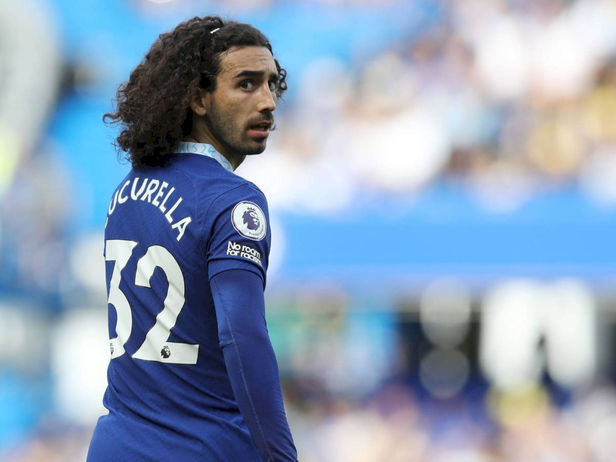 Football Players 桌布 壁紙 — 一位捲髮足球員身穿藍色球衣，在陽光明媚的球場上轉頭越過肩膀。