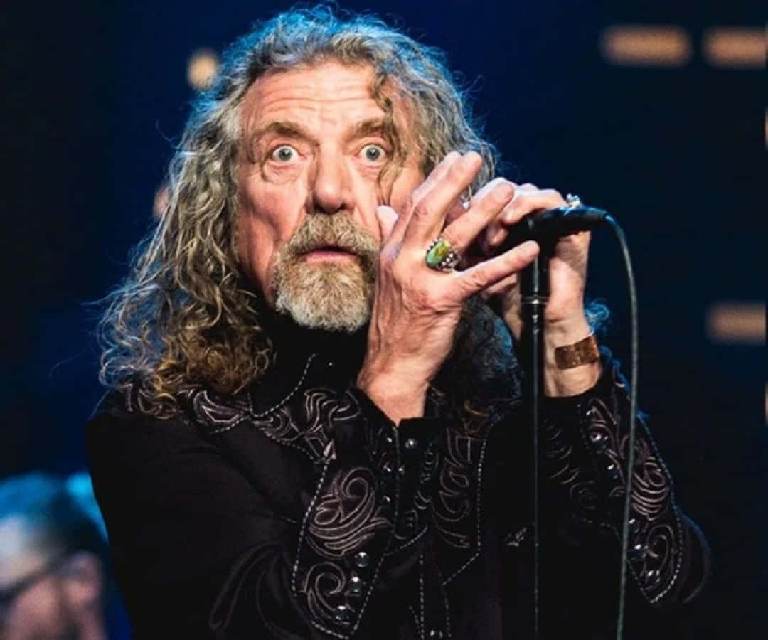 robert plant 桌布 壁紙 — 一位搖滾歌手，有著狂野的捲髮和鬍鬚，睜大眼睛，手指上戴著戒指，對著麥克風唱歌。