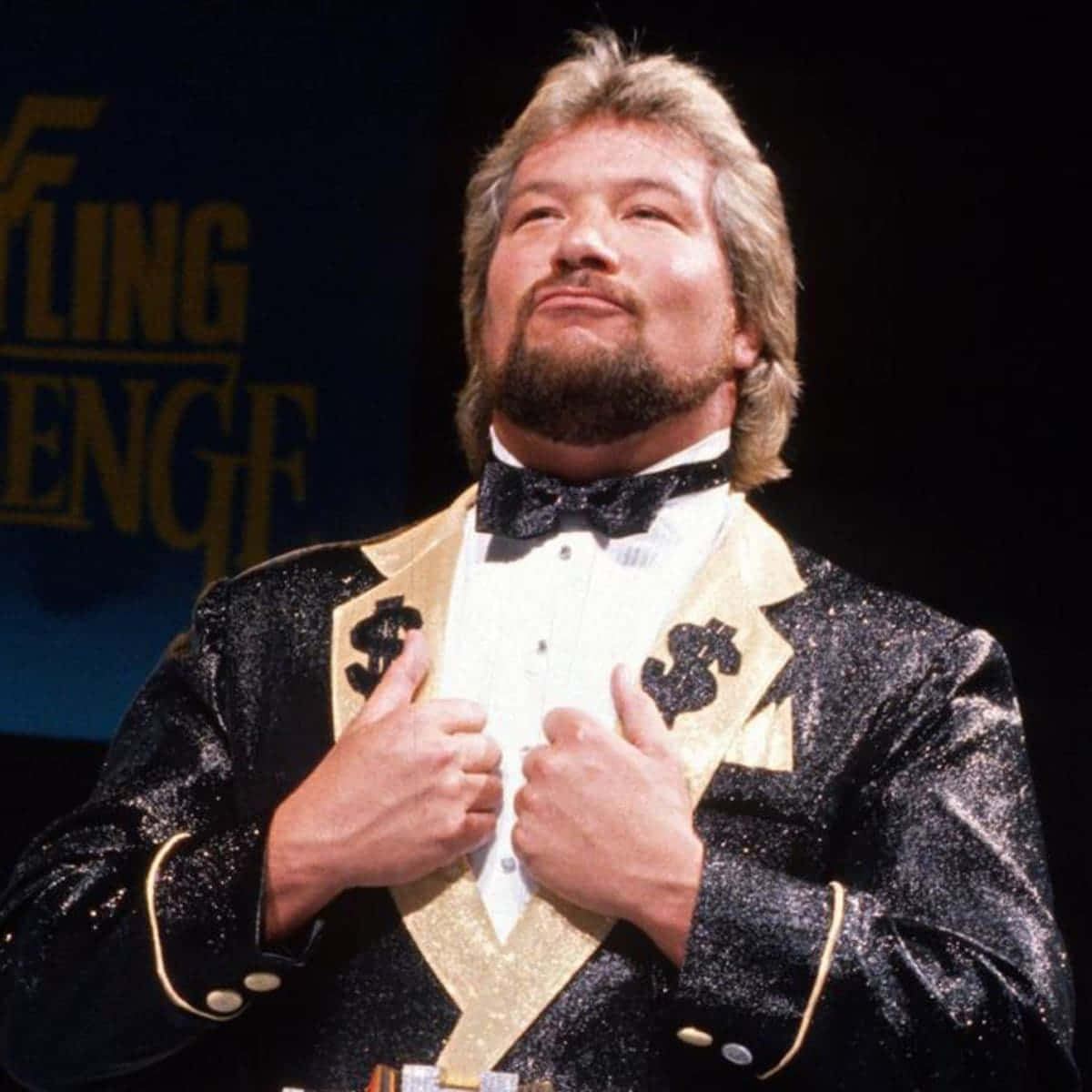 WWE 桌布 壁紙 — 一位摔角手身穿閃亮黑色金色西裝，領口有美元符號，自信地在舞台上擺姿勢。