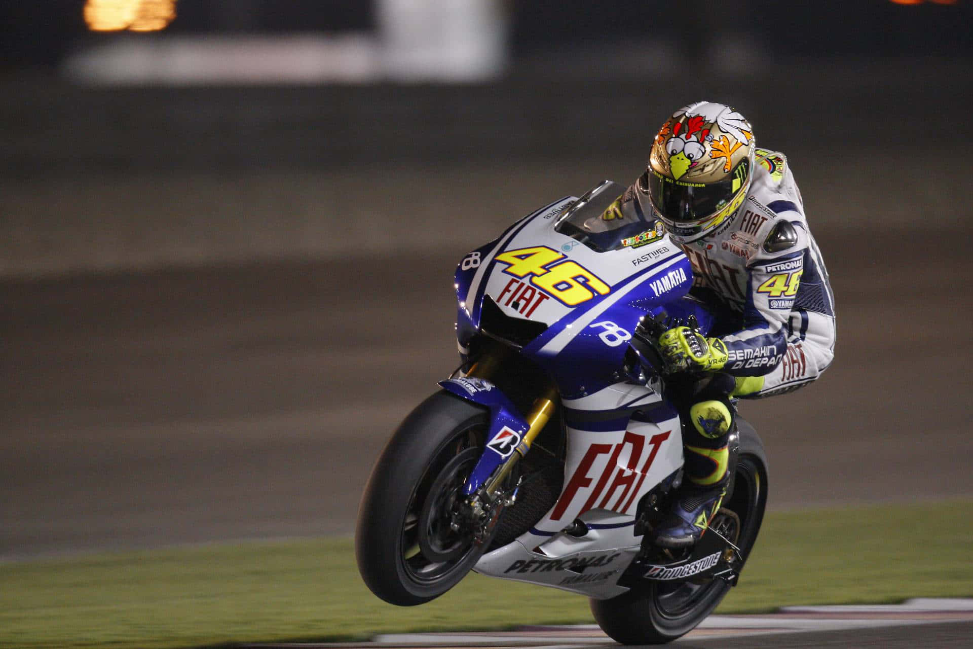 Motorsport 桌布 壁紙 — 一位摩托車手在夜間燈火通明的賽道上傾斜入彎，戴著色彩繽紛的安全帽和賽車服。