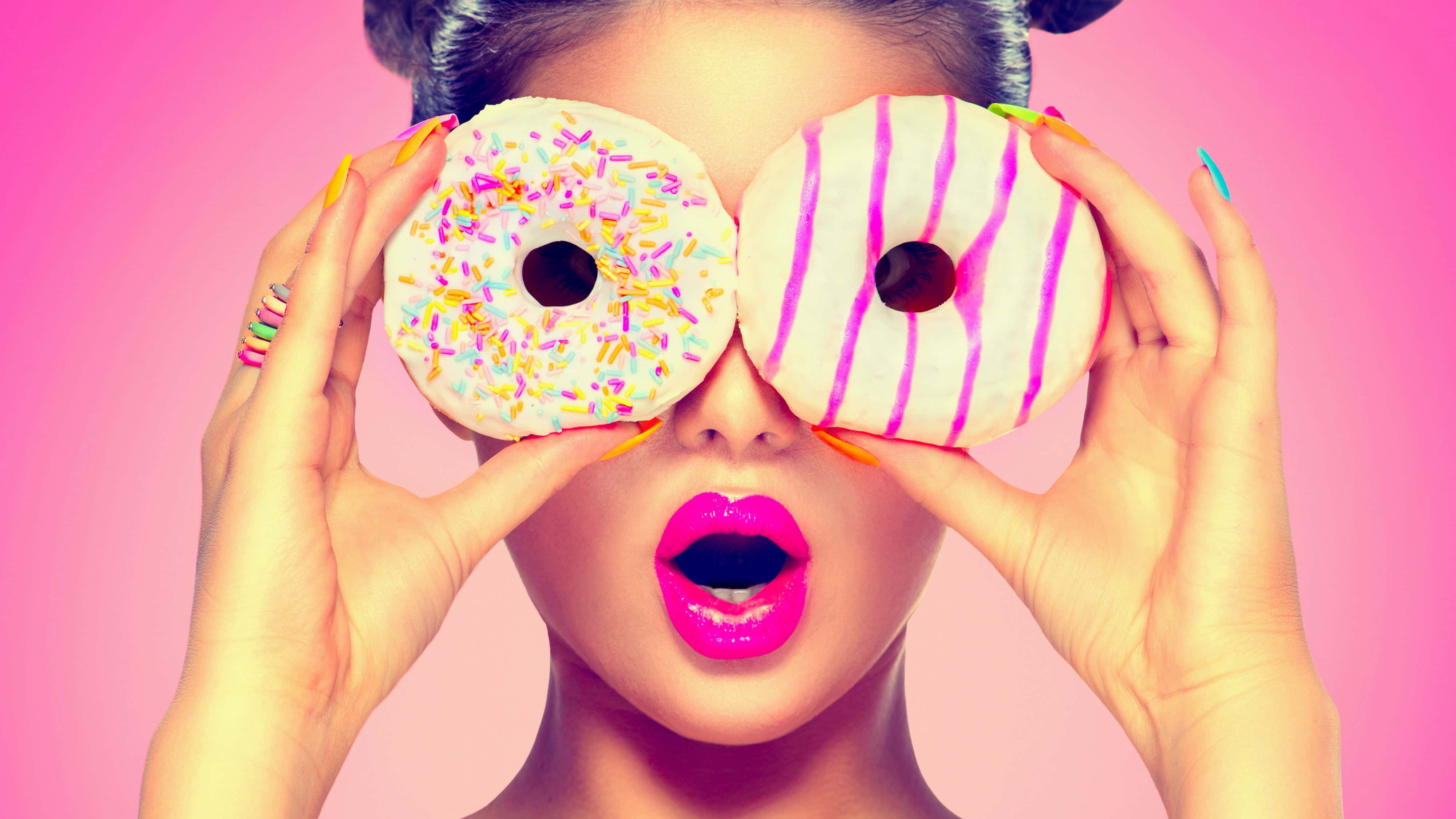 donuts 桌布 壁紙 — 一位擁有亮粉紅色嘴唇的女性，在鮮豔的粉紅色背景前，將兩個裝飾過的甜甜圈舉在眼前。