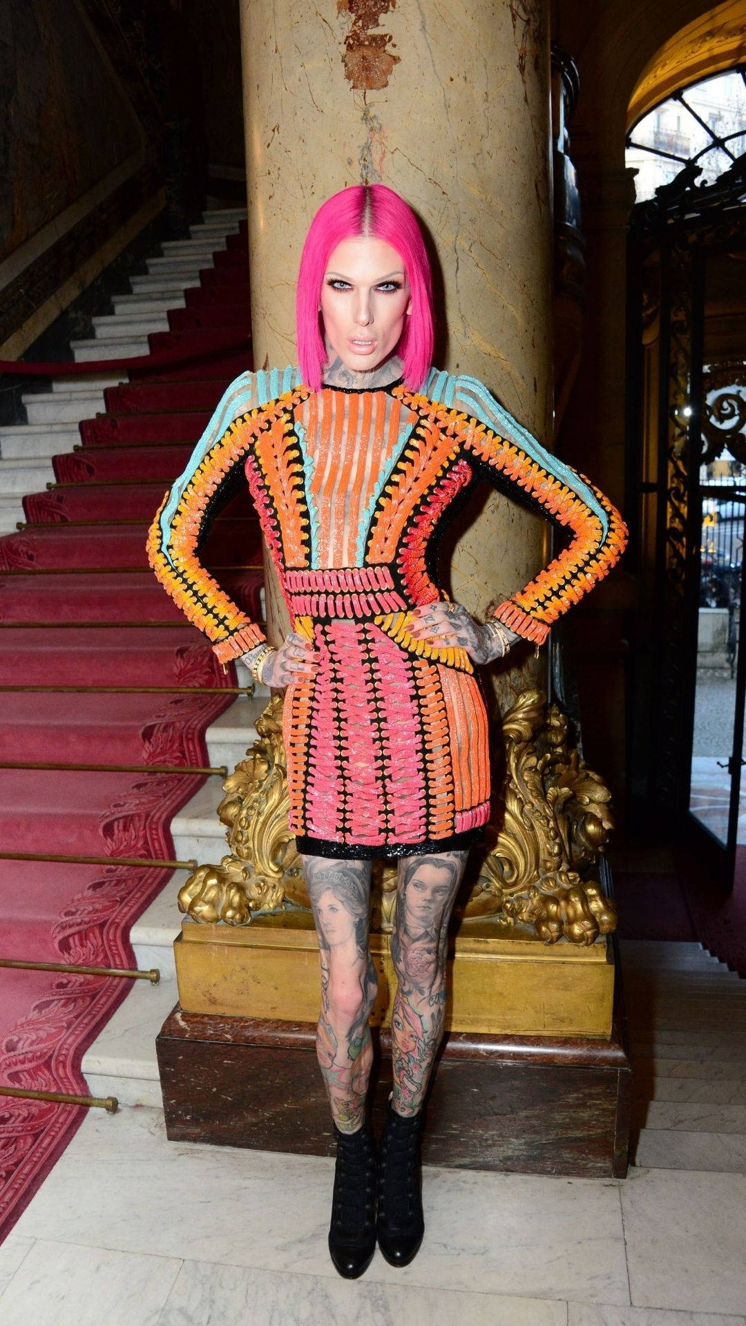 Jeffree Star 桌布 壁紙 — 一位擁有亮粉紅色頭髮和刺青的女性，穿著色彩繽紛的幾何圖案連衣裙，自信地在大樓梯旁擺姿勢。