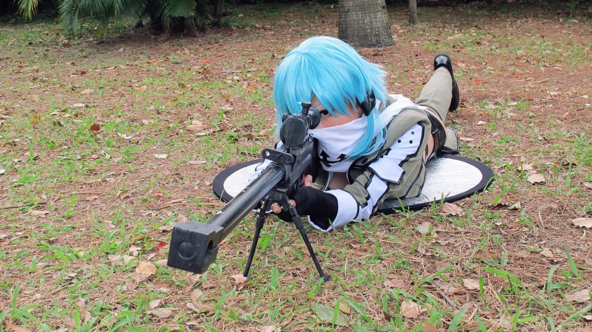 Sword Art Online Cosplay 桌布 壁紙 — 一位擁有亮藍色頭髮和白色面具的角色扮演者，俯臥在草地公園中，瞄準一支帶瞄準鏡的大型黑色步槍。